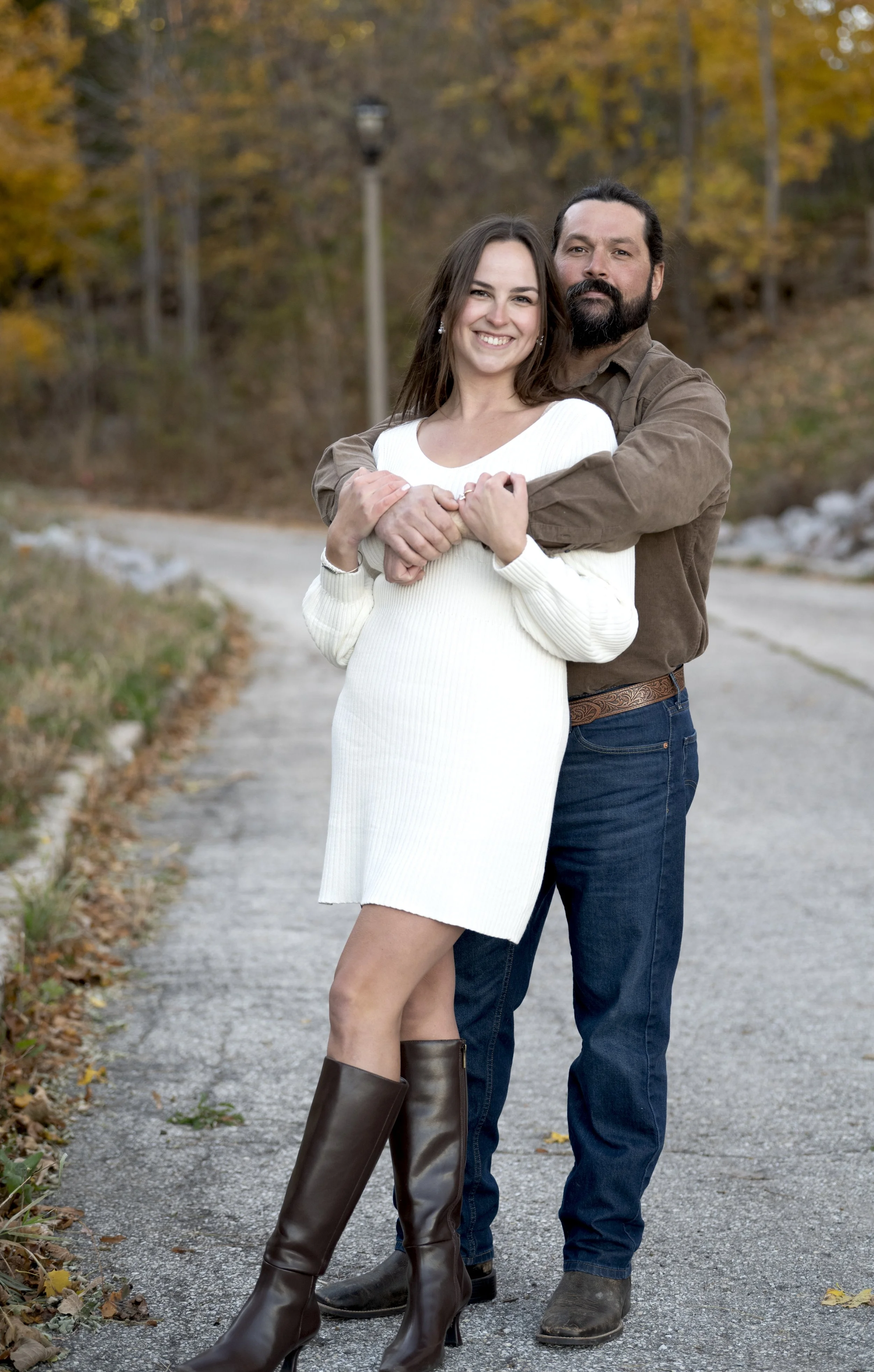 A smiling woman wearing a white sweater dress and knee-high brown boots is hugging a man with a beard, who is wearing a brown shirt and jeans, on a gravel path outdoors with autumn trees in the background.