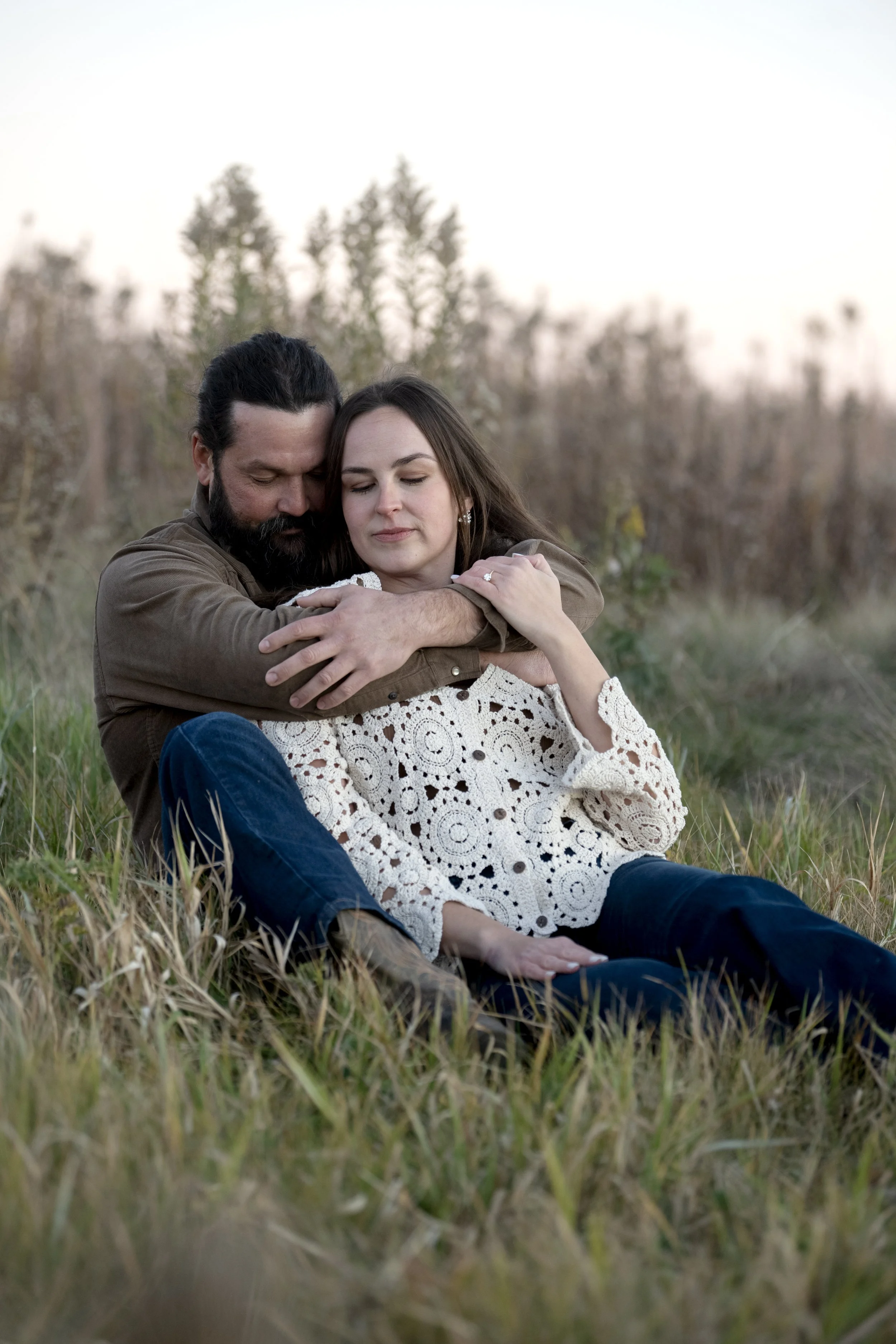 A man and a woman sitting in a grassy field, embracing each other with their eyes closed, surrounded by blurred trees and a soft sky in the background.