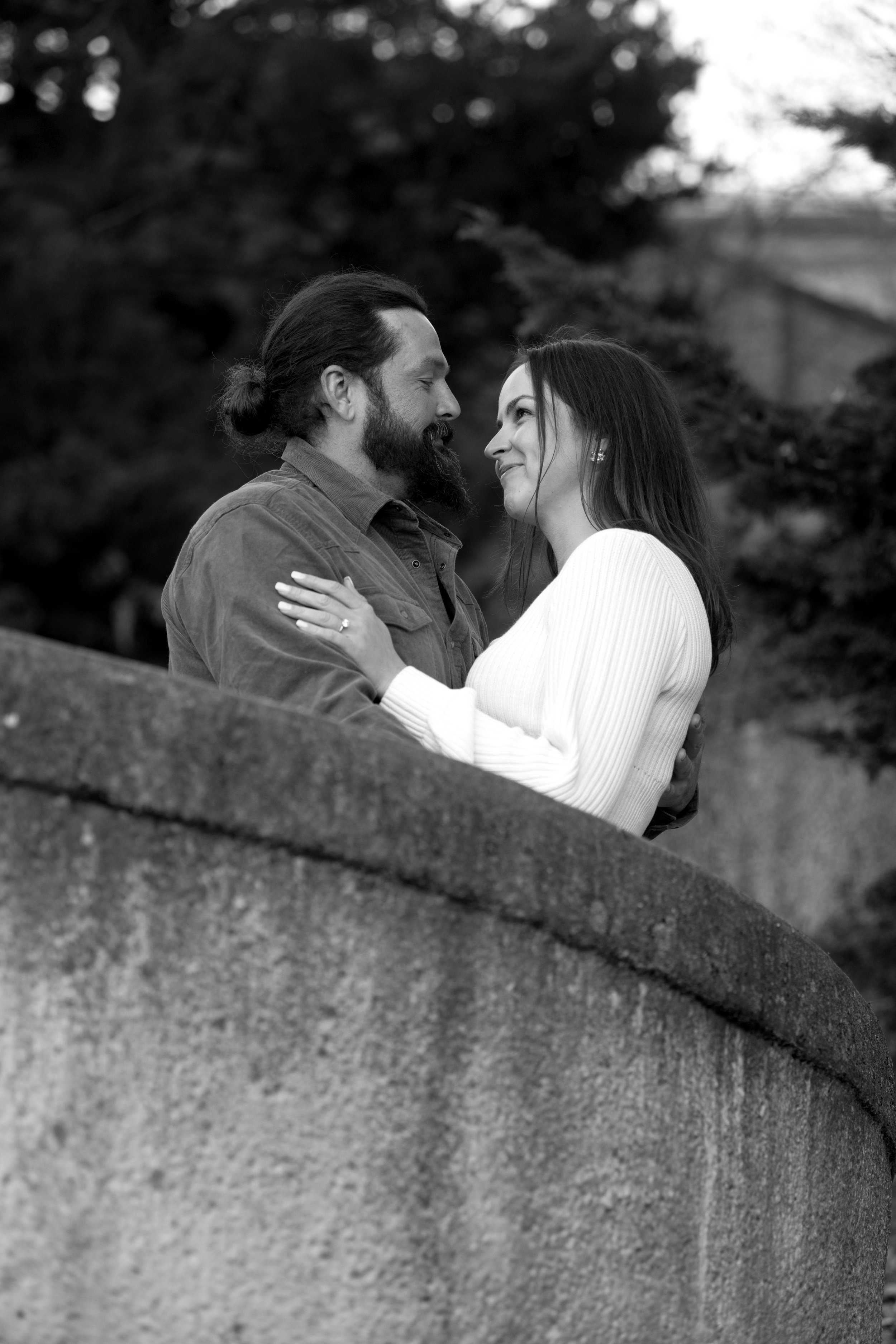 A black and white photo of a man with a beard and long hair tied back, and a woman with long dark hair, smiling at each other while embracing outdoors with trees in the background.