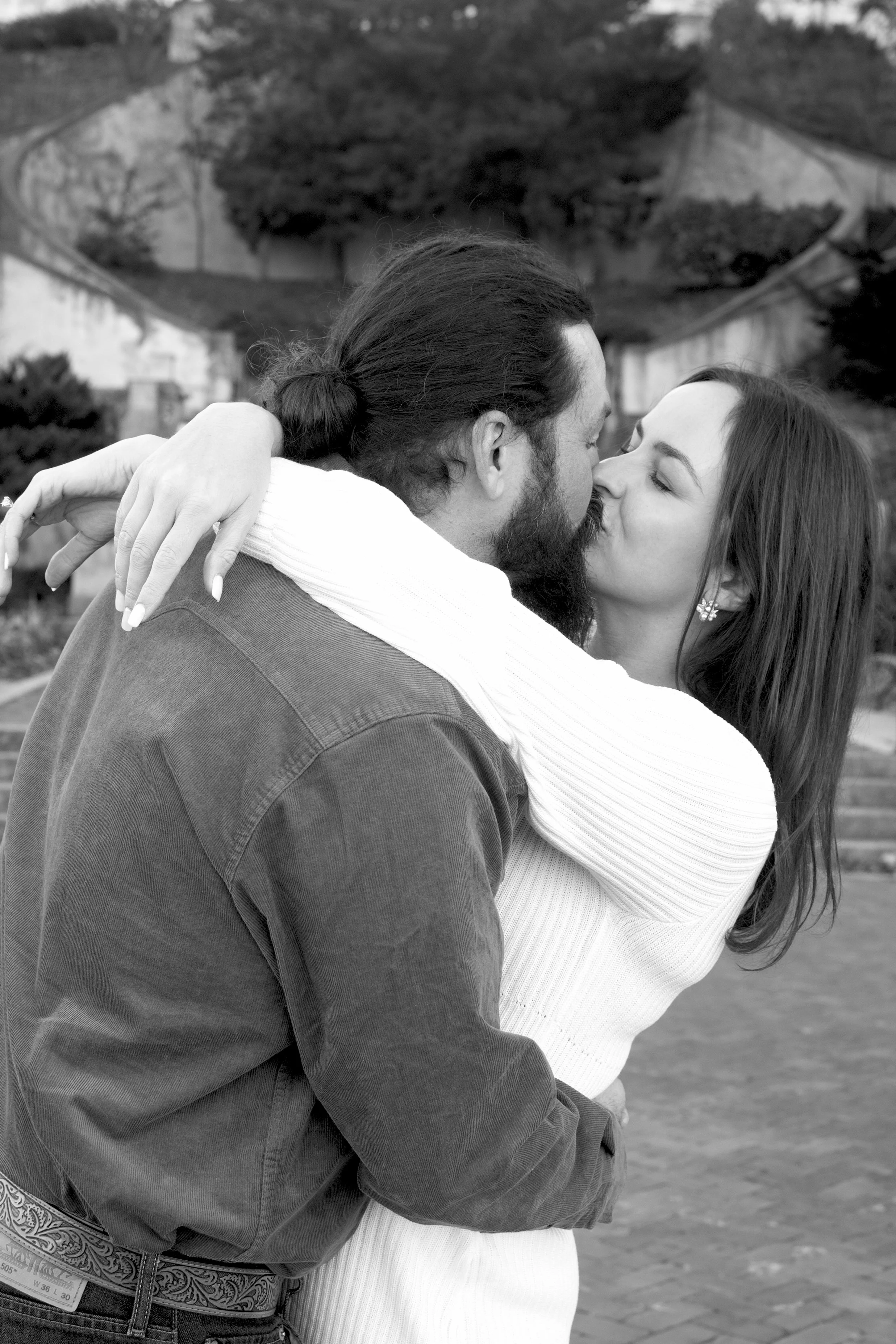 A black and white photo of a man and woman sharing a kiss outdoors. The man has long hair tied back and a beard, and the woman has long hair and is wearing earrings. They are embracing with the woman's arm wrapped around the man's neck.