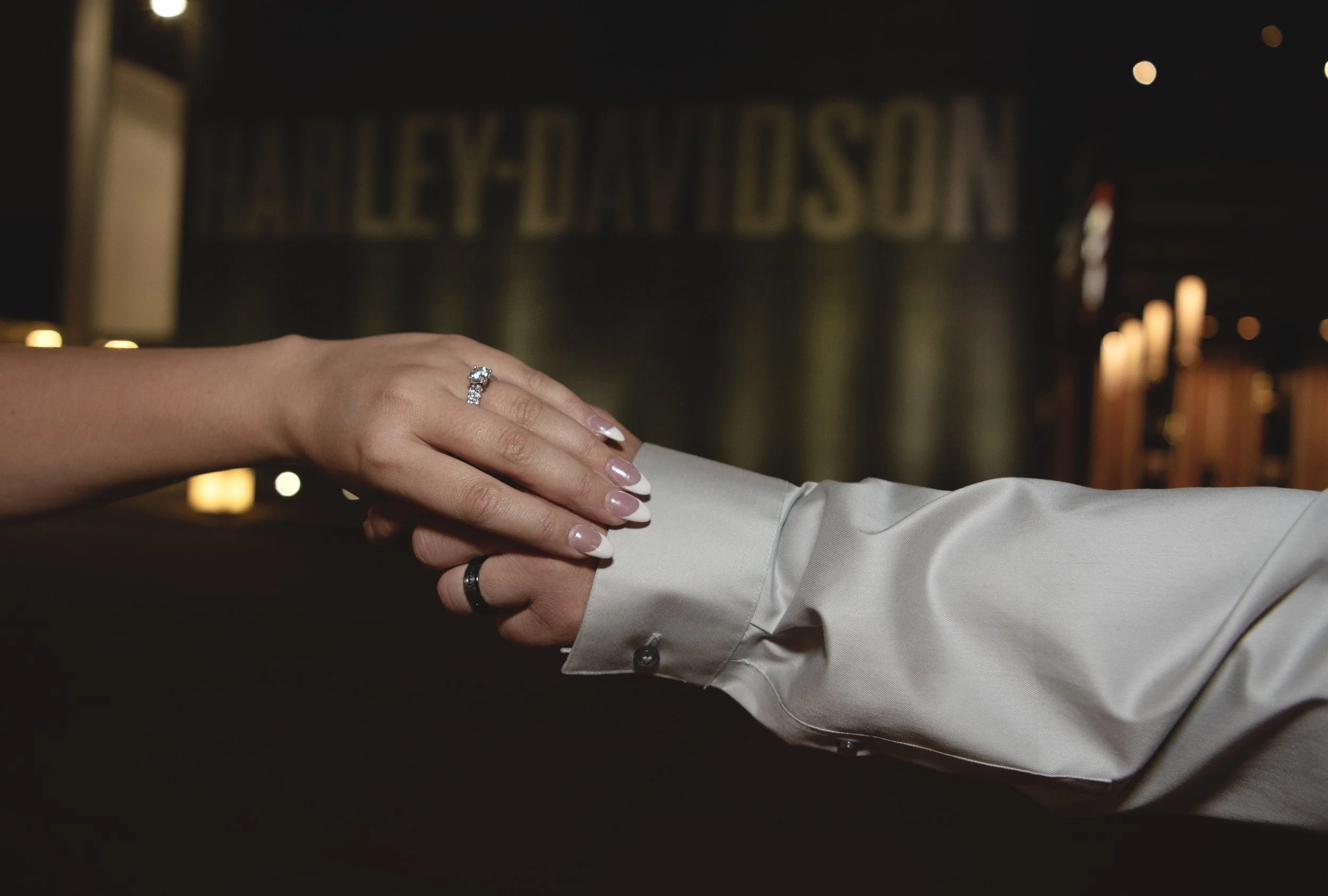 A woman's hand with a diamond engagement ring holds a man's hand. They are dressed in formal attire, and the background has blurred warm lighting and the word 'HENRY'S'.