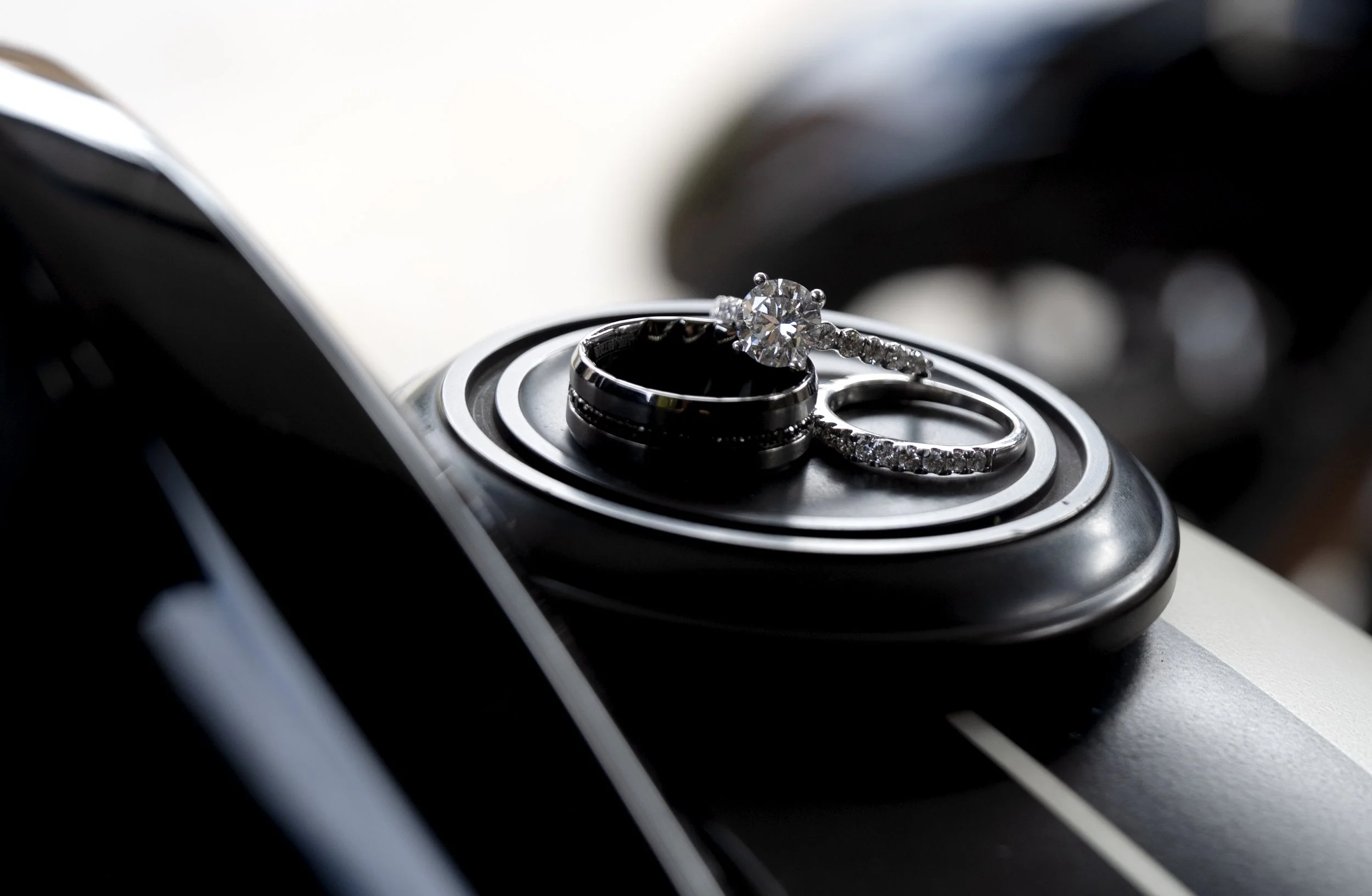 Close-up of a diamond engagement ring and a wedding band resting on a metallic part, possibly of a motorcycle or a similar object.