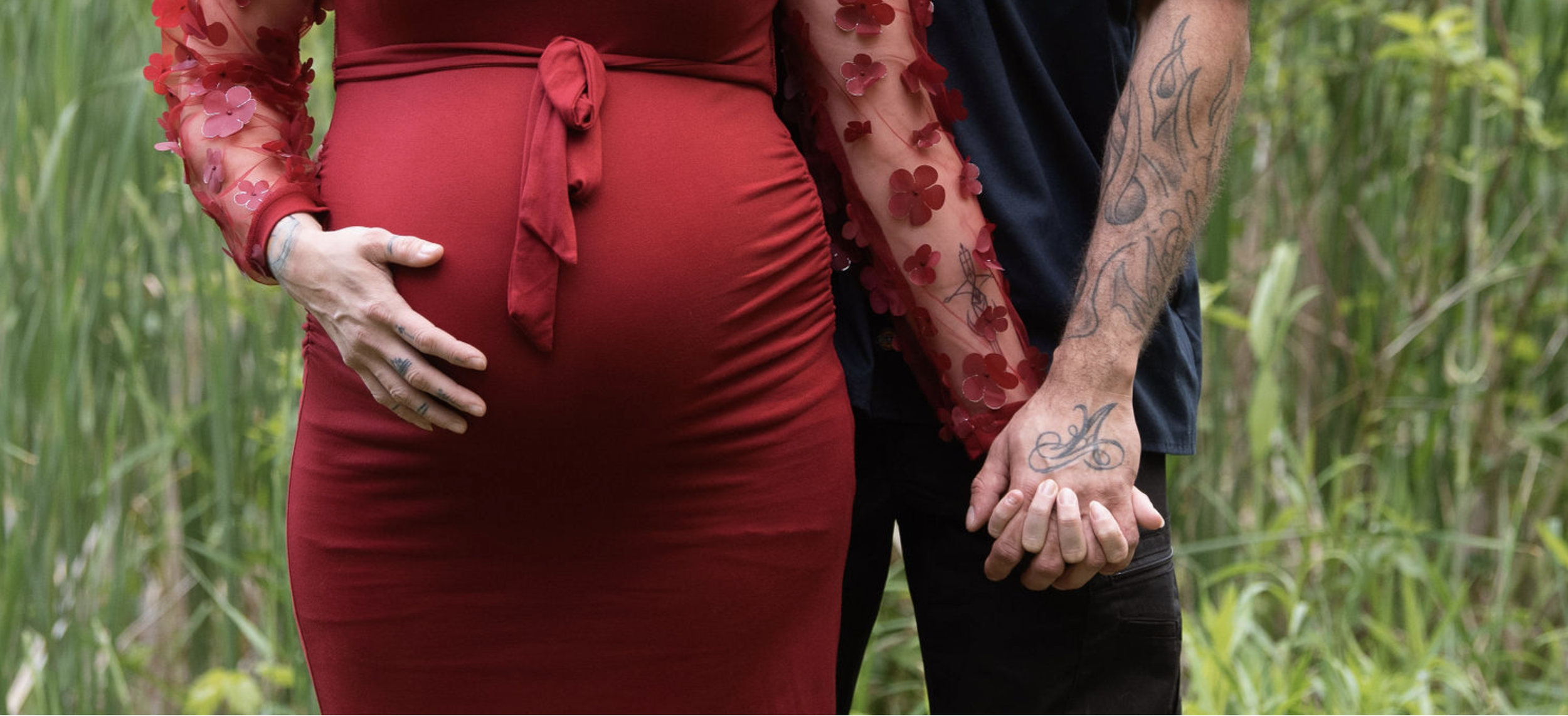 A pregnant woman wearing a red dress and a sheer red top with flower patterns holding hands with a man with tattoos, standing in a green outdoor setting.
