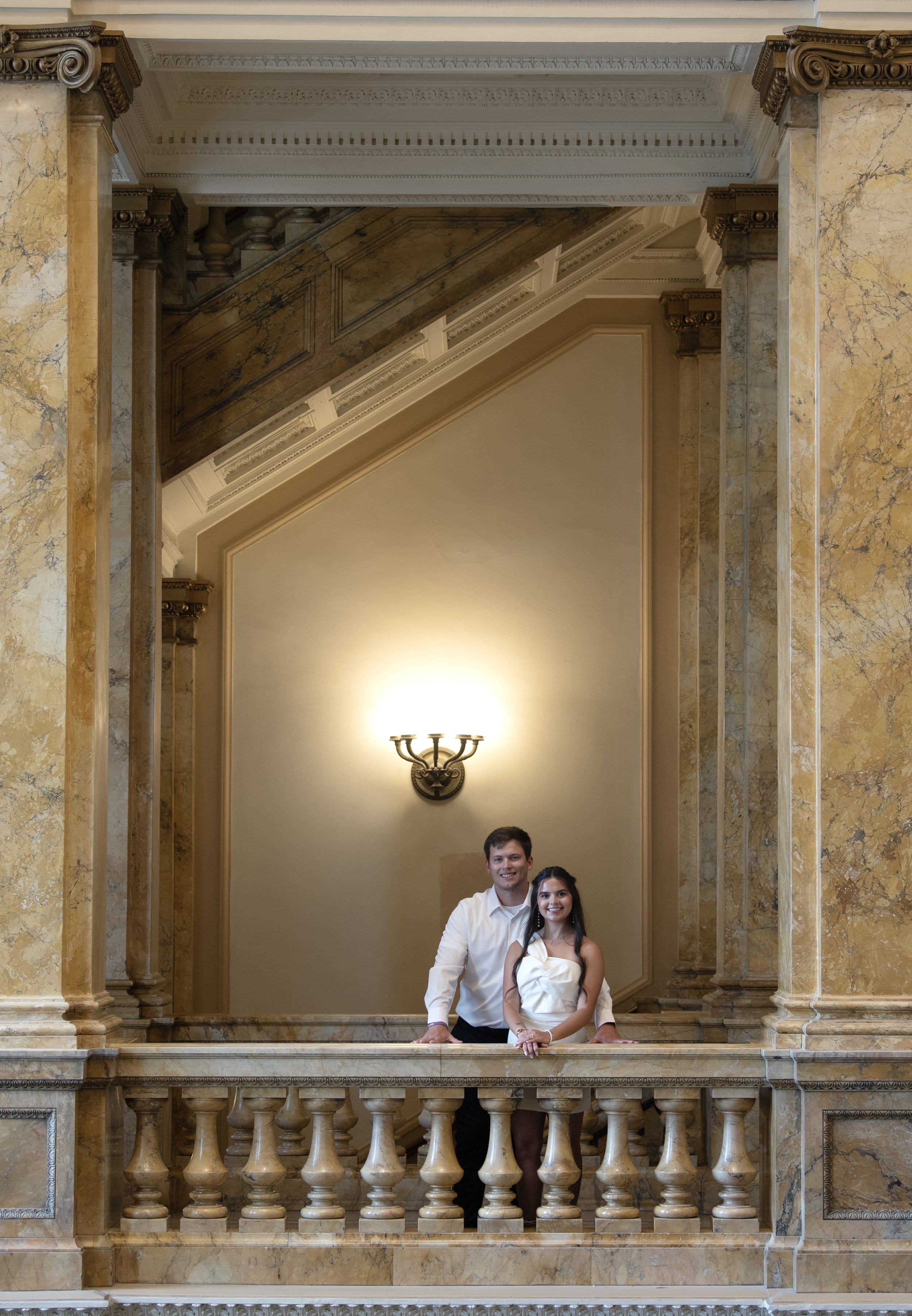 A smiling young man and woman standing on a marble balcony in an ornate, classical-style interior with columns, decorative moldings, and a chandelier.