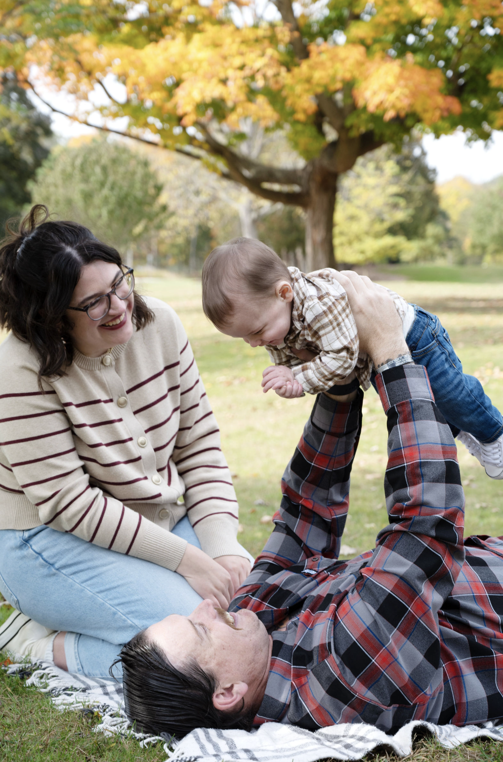 Family playing outdoors in a park with colorful fall trees. A man lying on a blanket lifts a smiling toddler while a woman watches and laughs.