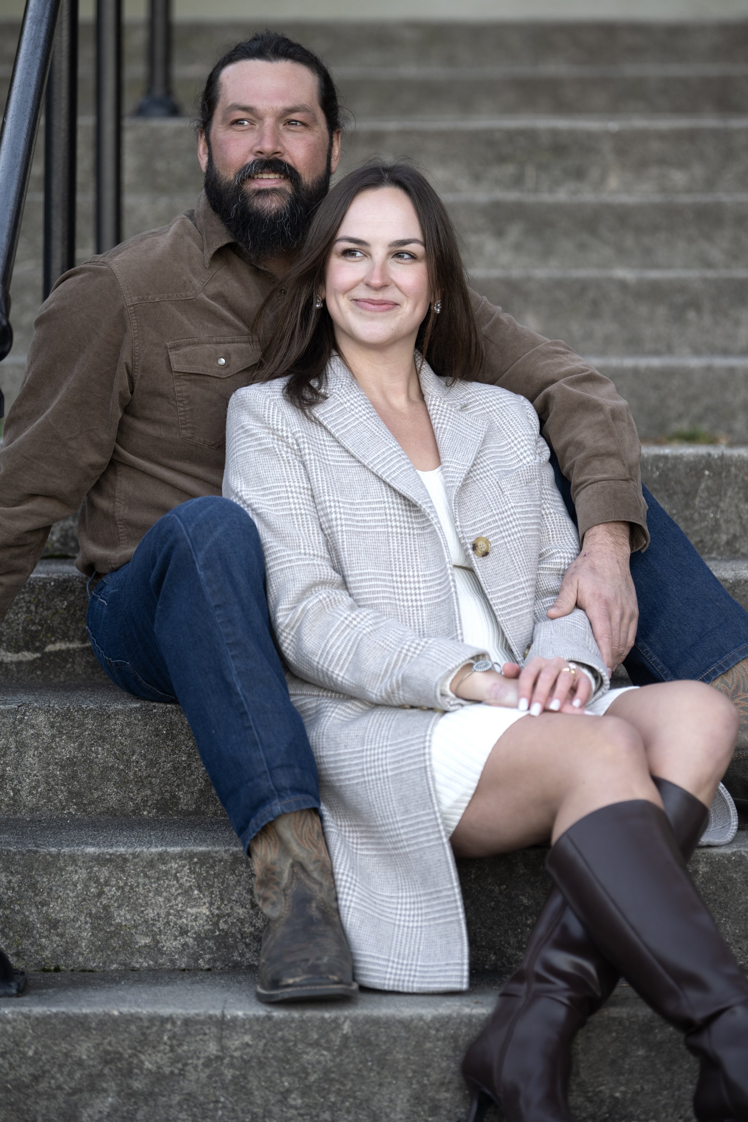 A man and a woman sitting on outdoor steps. The man has a beard, long dark hair, and is wearing a brown shirt. The woman has shoulder-length dark hair, earrings, a beige plaid blazer, and a white dress. They are smiling and looking to the side.