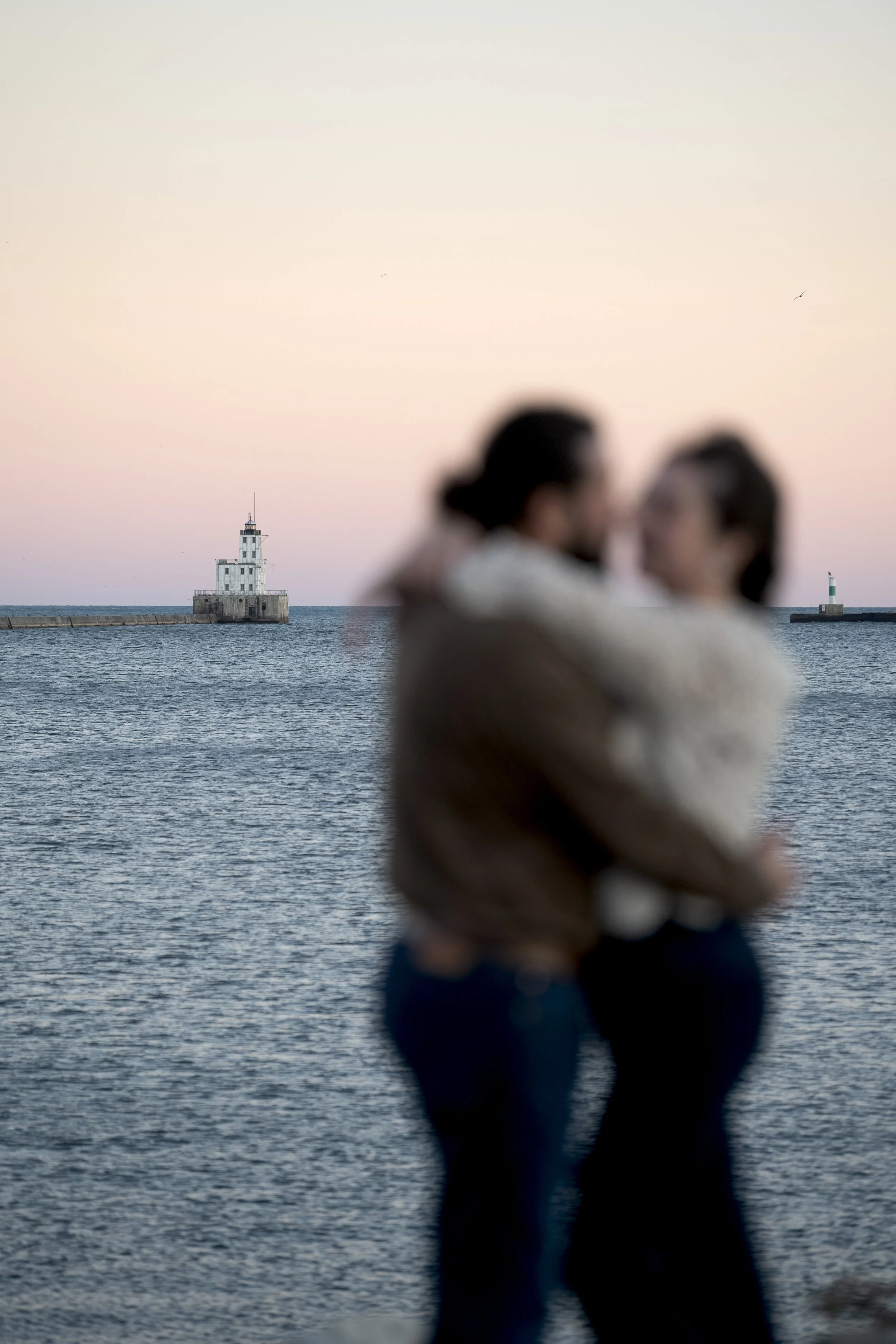 Two blurred people standing near the water with a lighthouse in the background at sunset.