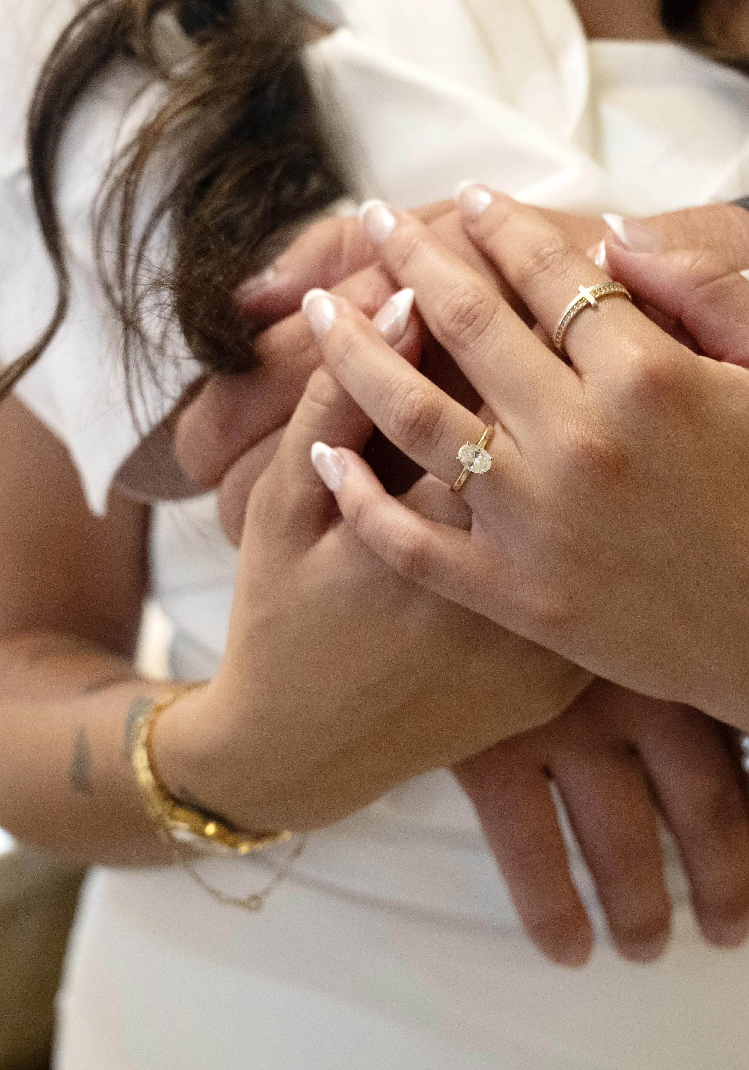 Close-up of two women holding hands, showing jewelry including rings and bracelets, with one woman having a French manicure and a tattoo.