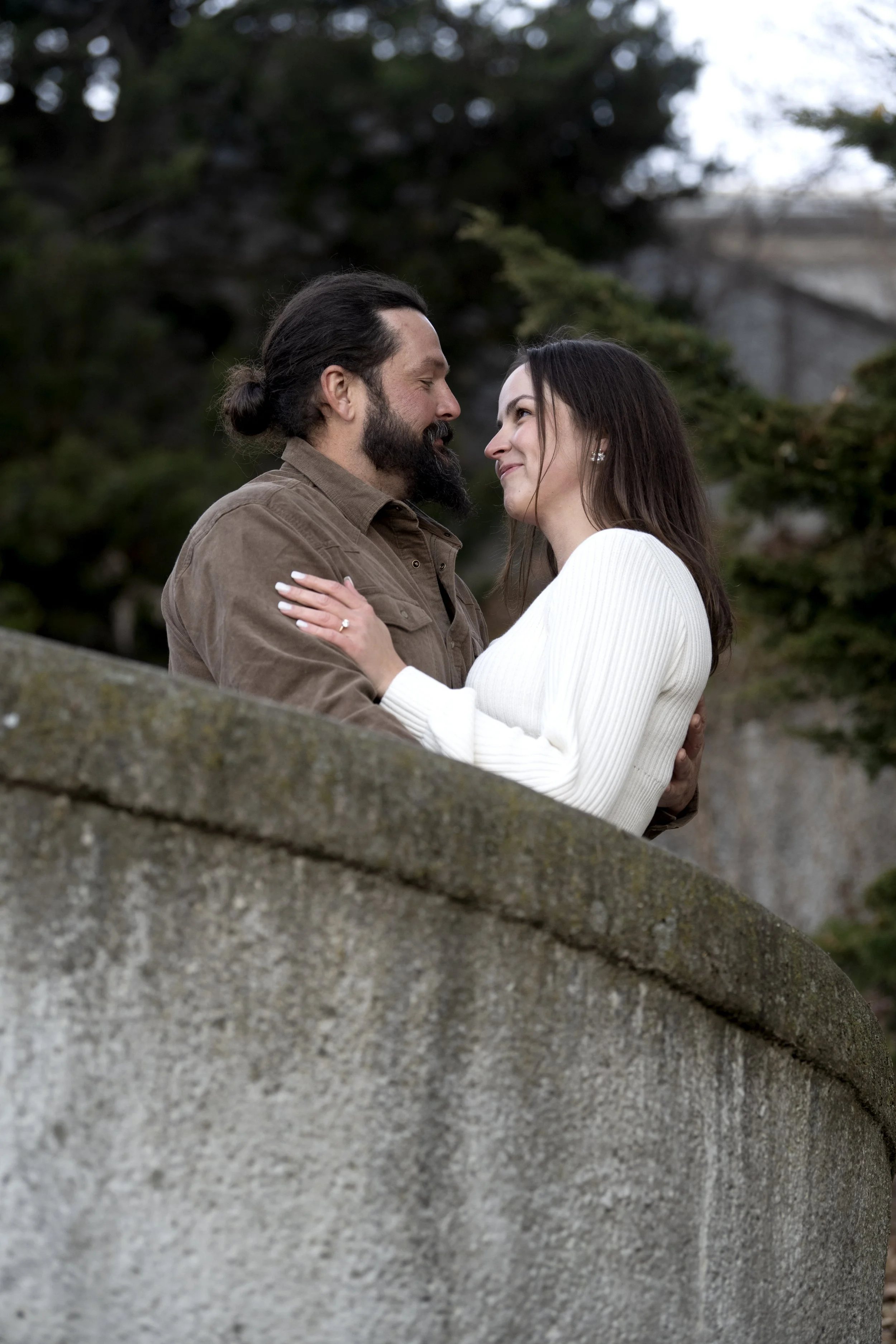 A man and a woman stand facing each other outdoors, with the man holding the woman's upper arms, both smiling and looking into each other's eyes, surrounded by trees.