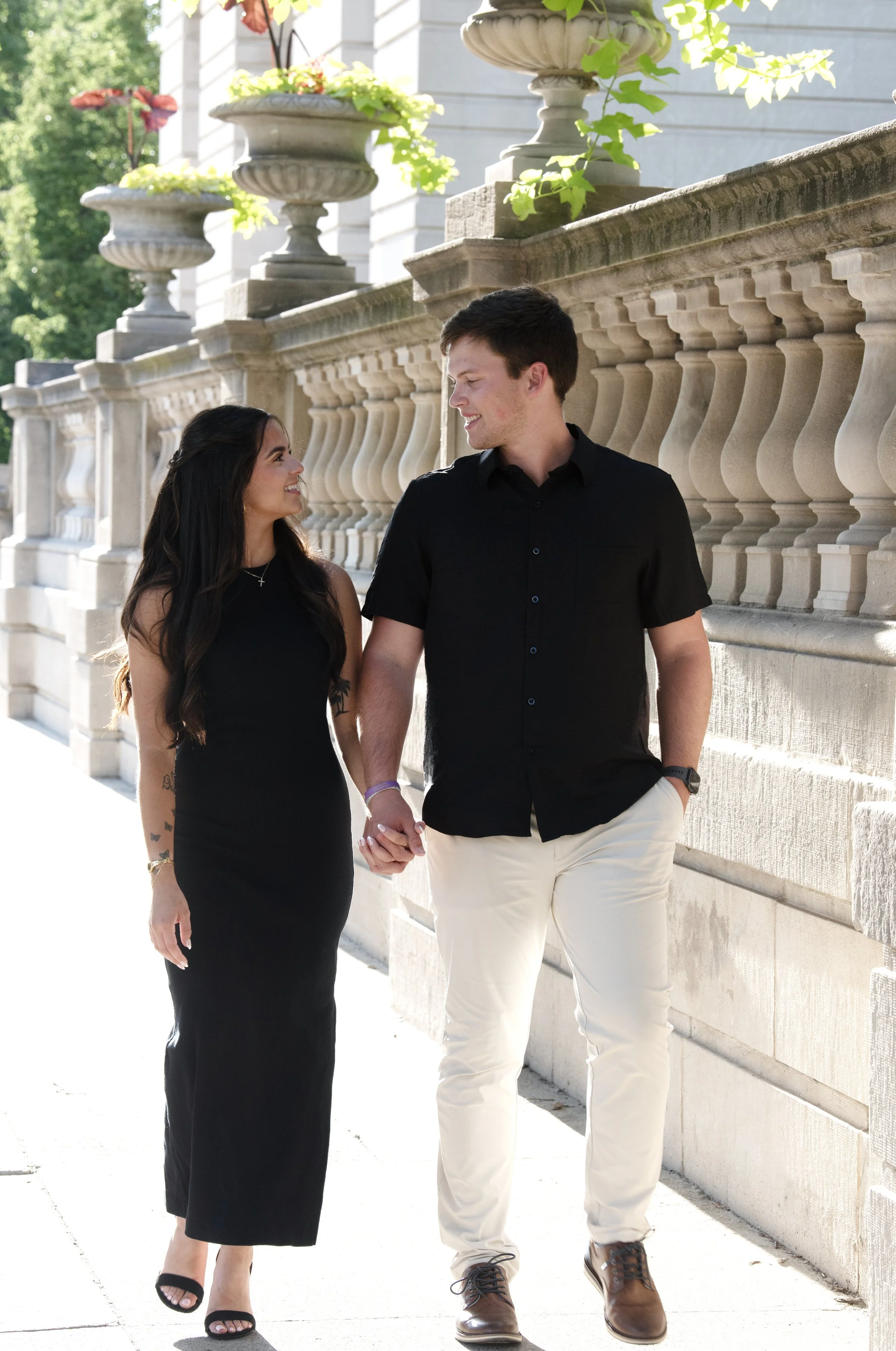 A young couple walking hand in hand outdoors, smiling at each other, with a stone railing and large decorative planters with green plants and flowers in the background.