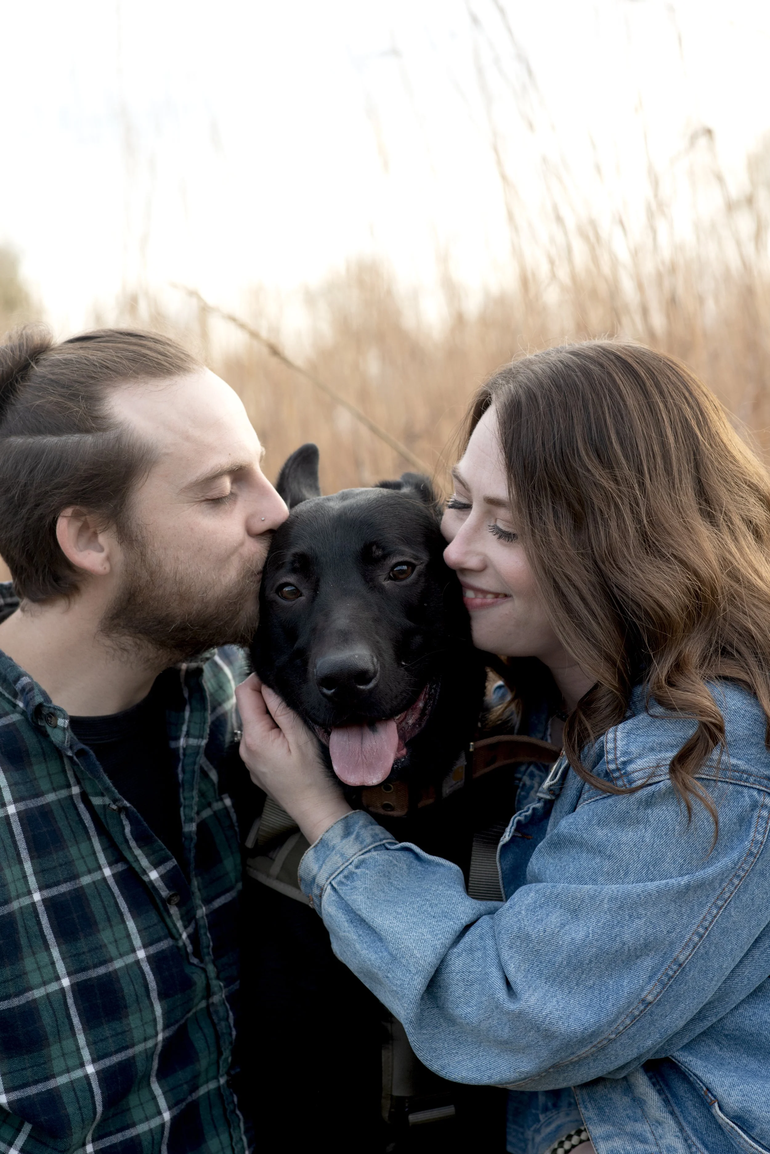 A man and a woman hugging a black dog with its tongue out, in an outdoor setting with tall, dry grass in the background.