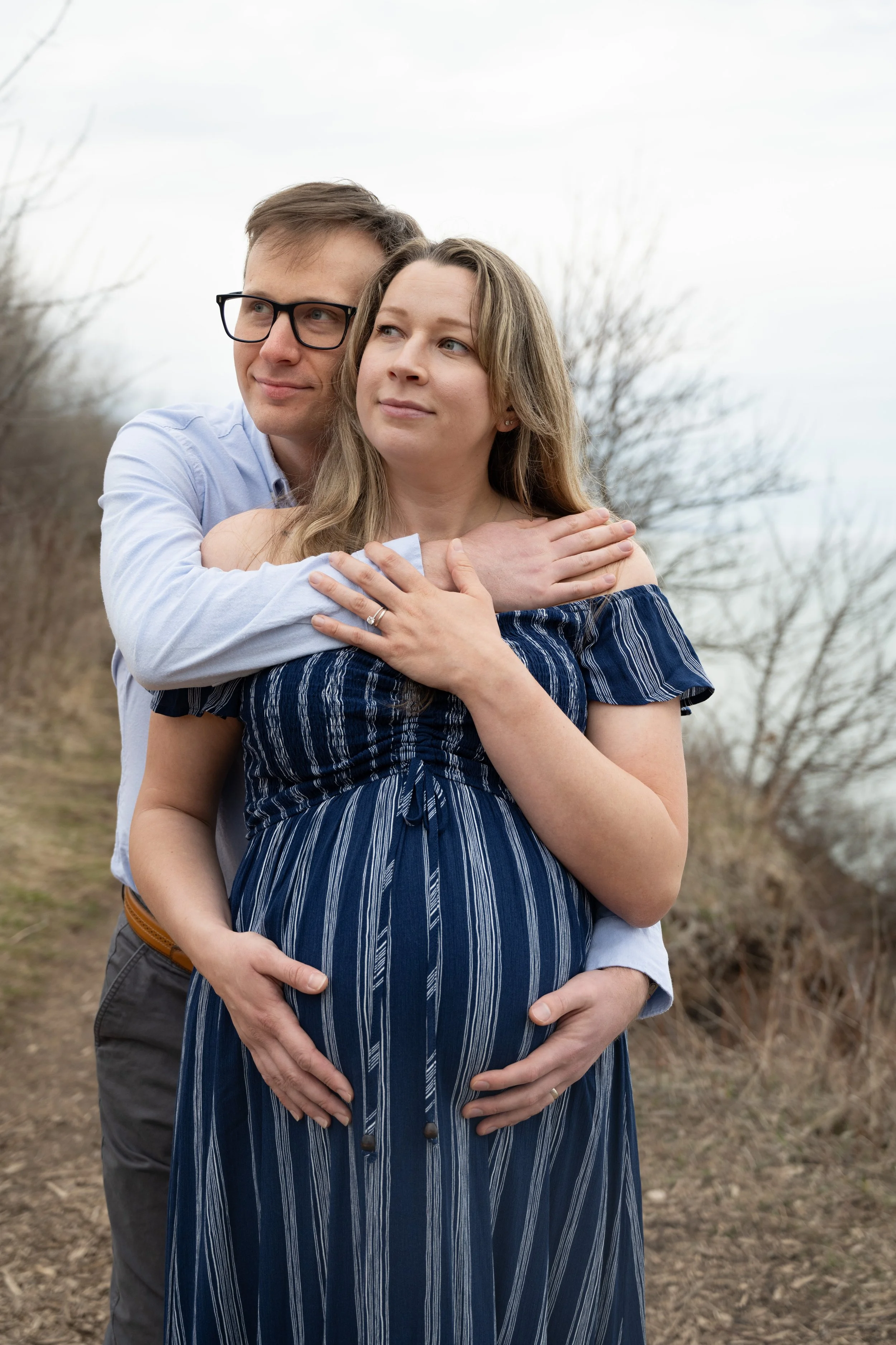 A couple standing outdoors, with the man embracing the pregnant woman from behind, both looking thoughtfully into the distance. The woman is wearing a blue and white striped dress, and the man is wearing glasses and a light blue shirt. The background includes leafless trees and a cloudy sky.