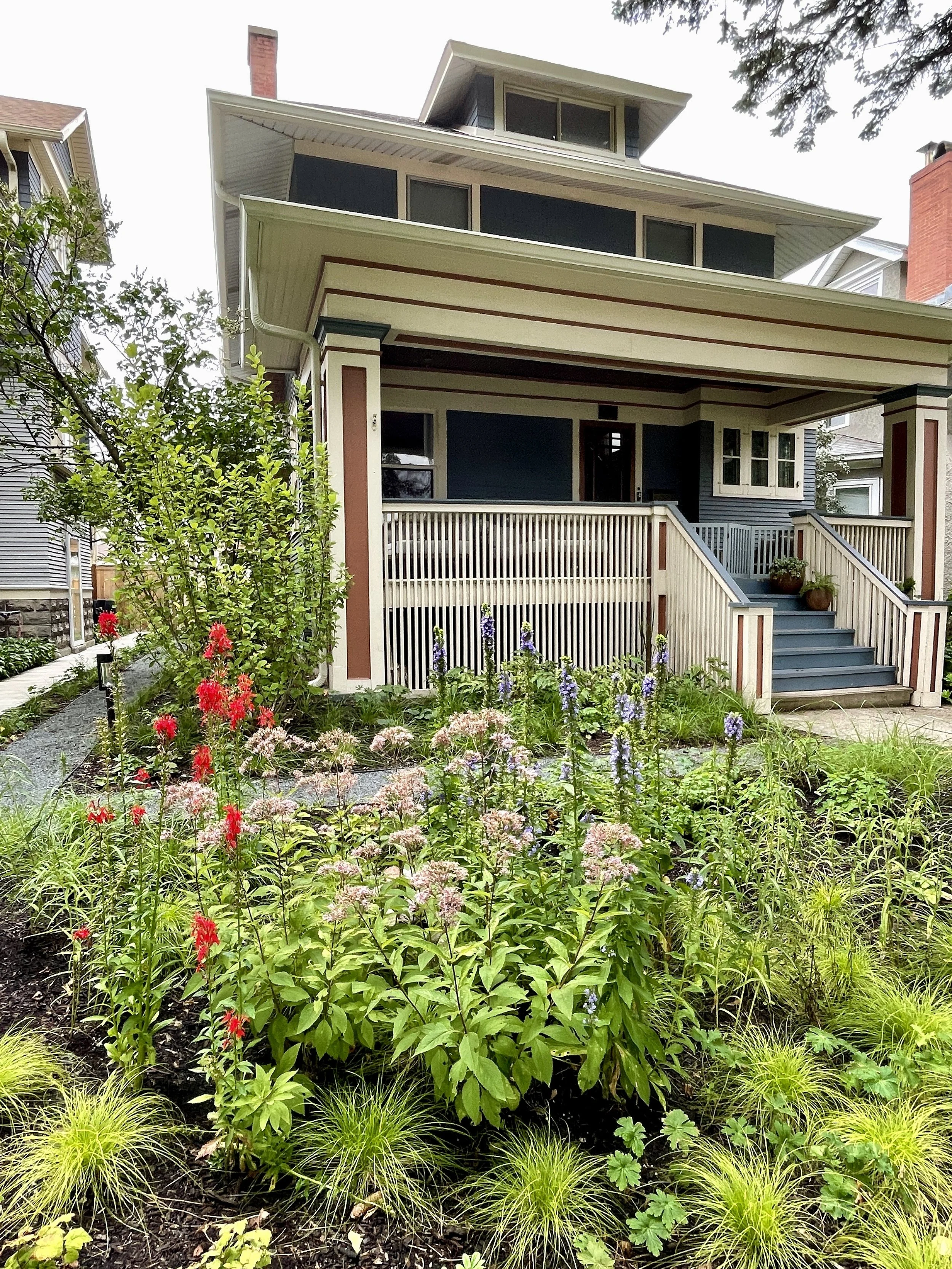 A two-story house with a porch, stairs, and a well-maintained garden with various green plants and purple, red, and pink flowers.