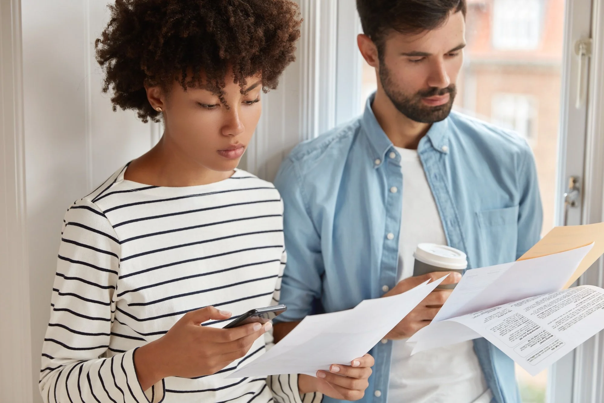 A woman with curly hair and a striped shirt looking at her phone, and a man with short dark hair and a beard wearing a light blue shirt reading papers and holding coffee, both standing by a window in a bright room.