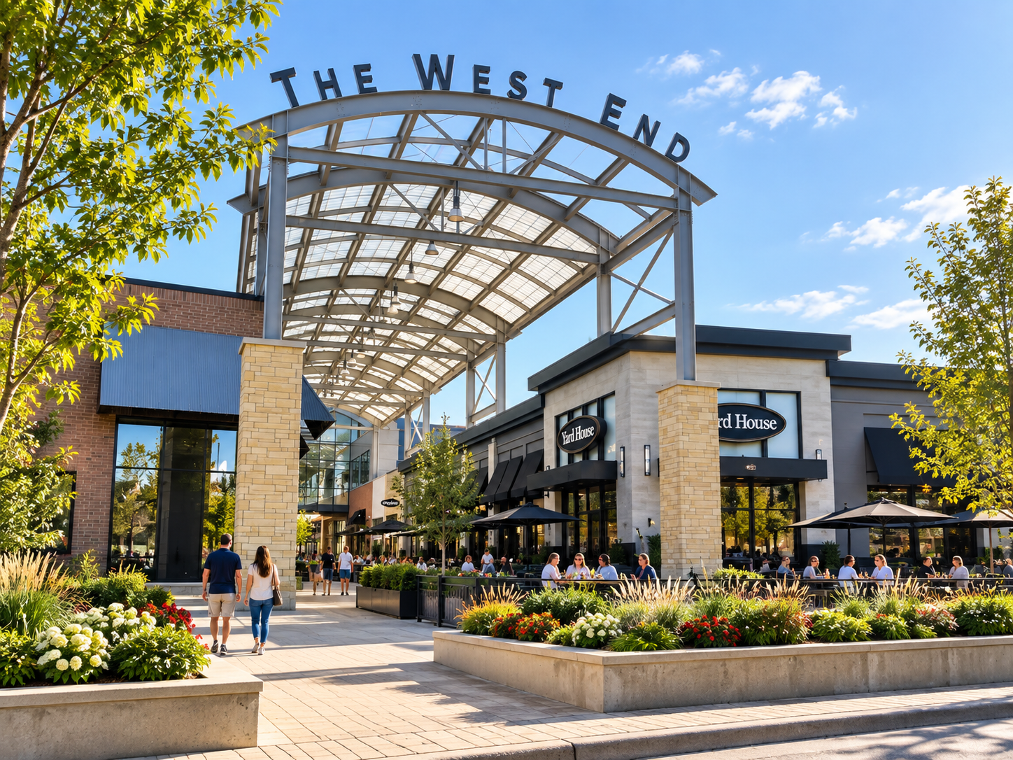 Outdoor shopping mall with storefronts, outdoor seating, people walking, and a large sign saying 'The West End' under a clear sky.
