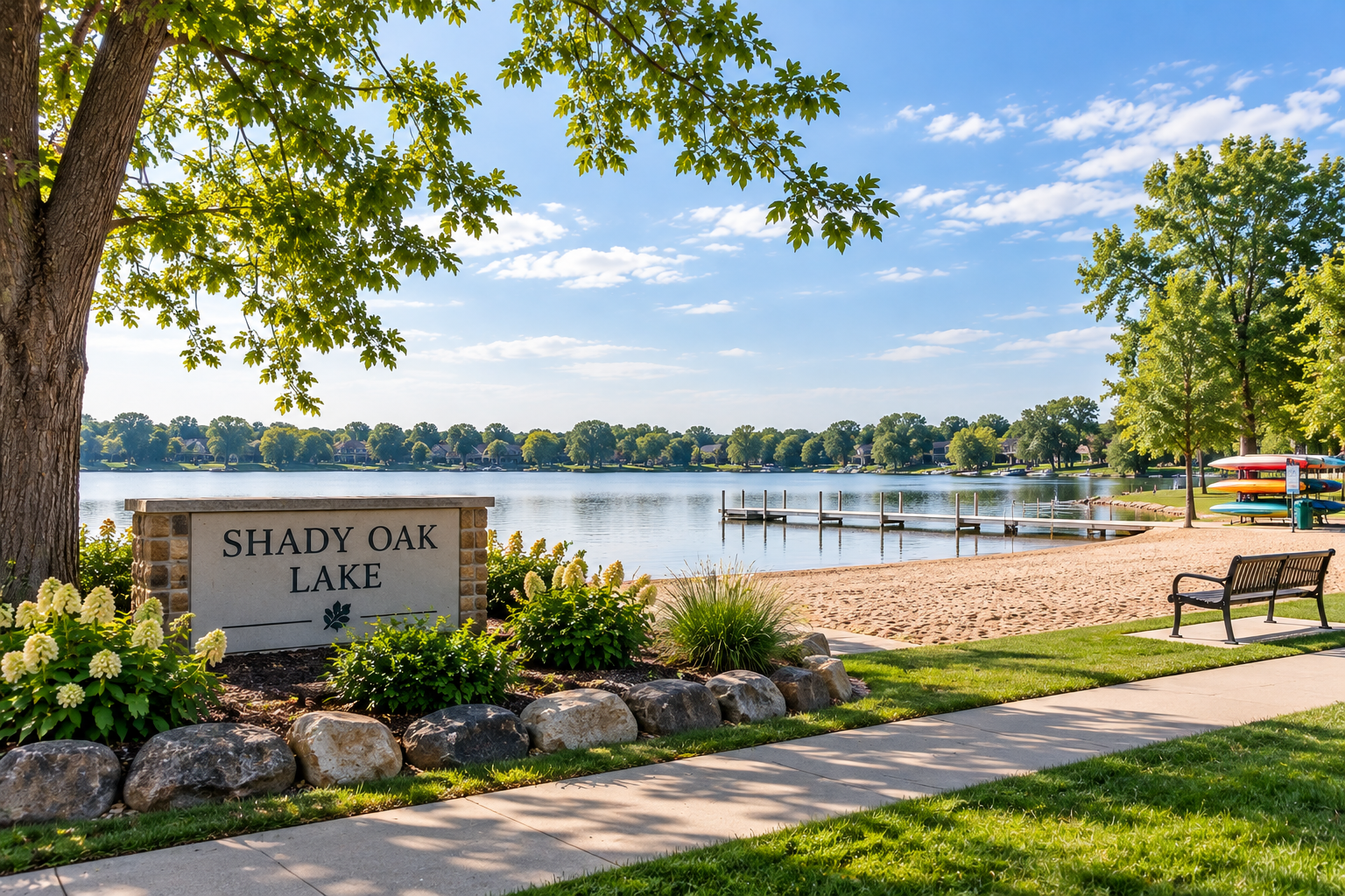 A peaceful lakeside scene at Shady Oak Lake showing a sandy beach, a dock stretching into the water, green trees, a bench, and colorful kayaks stacked near trees, with a partly cloudy blue sky