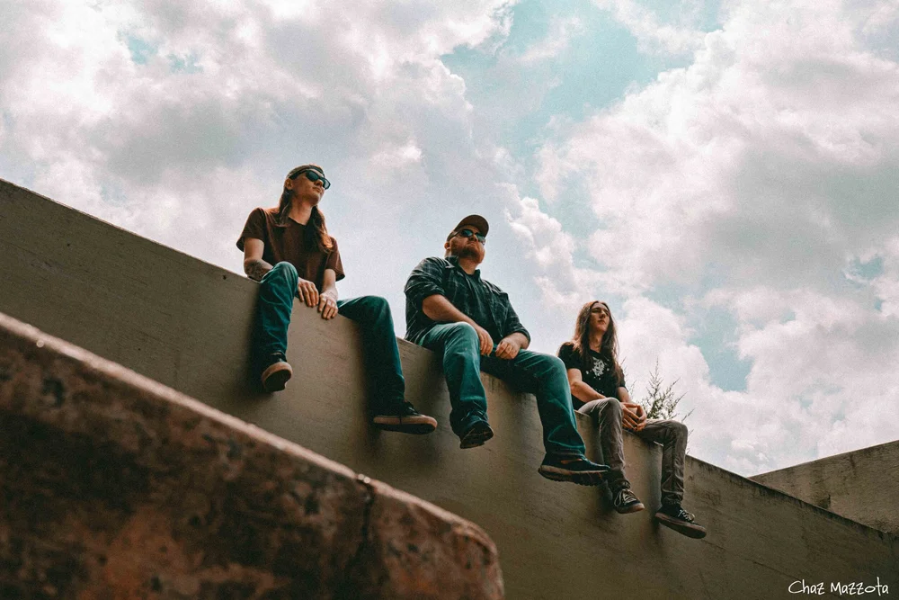 The band Sycamore sitting on a concrete wall under a cloudy sky, photographed from below by Chaz Mazzota