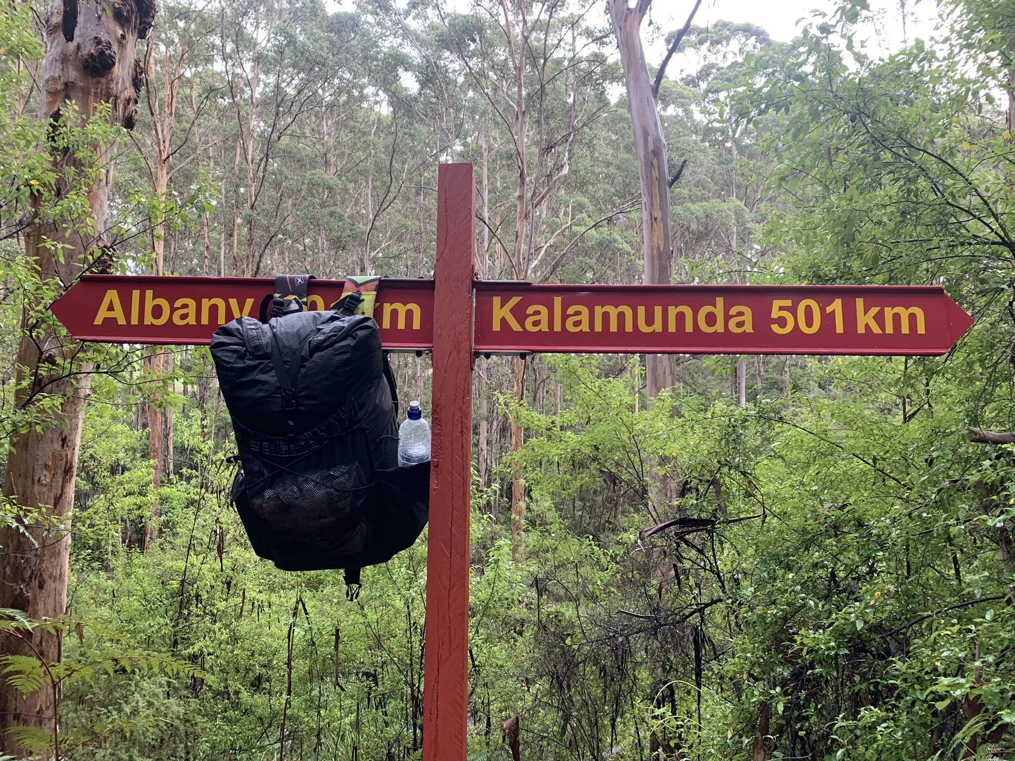 A directional sign at the Bibbulmun halfway point, in a forest indicating 501 kilometers to Kalamunda and 501 km to Albany, with a hiking backpack and water bottle hanging from the signpost.