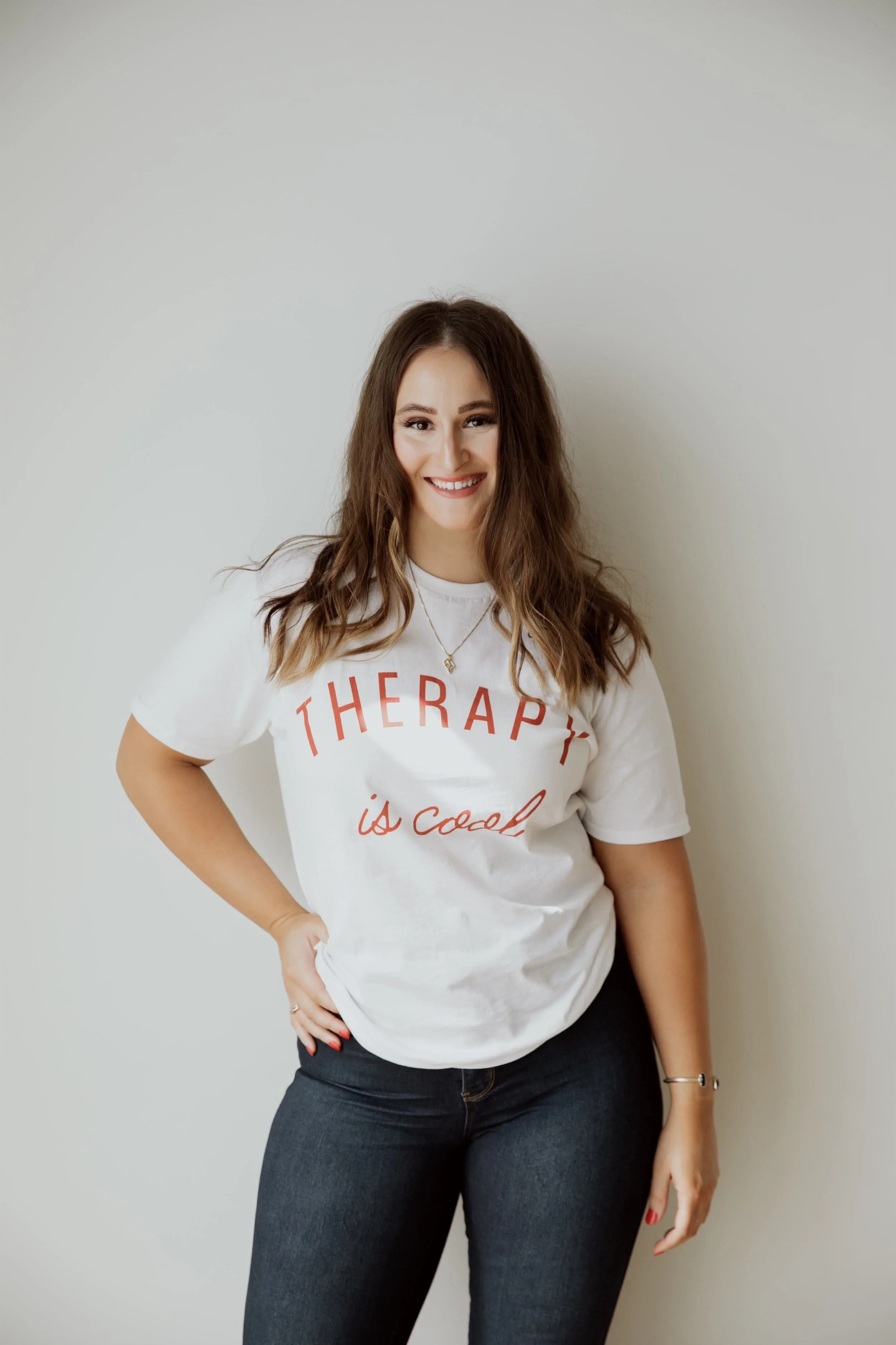 A woman with long wavy brown hair smiling, wearing a white t-shirt with 'THERAPY is cool' written on it, and black jeans, standing against a plain white wall.