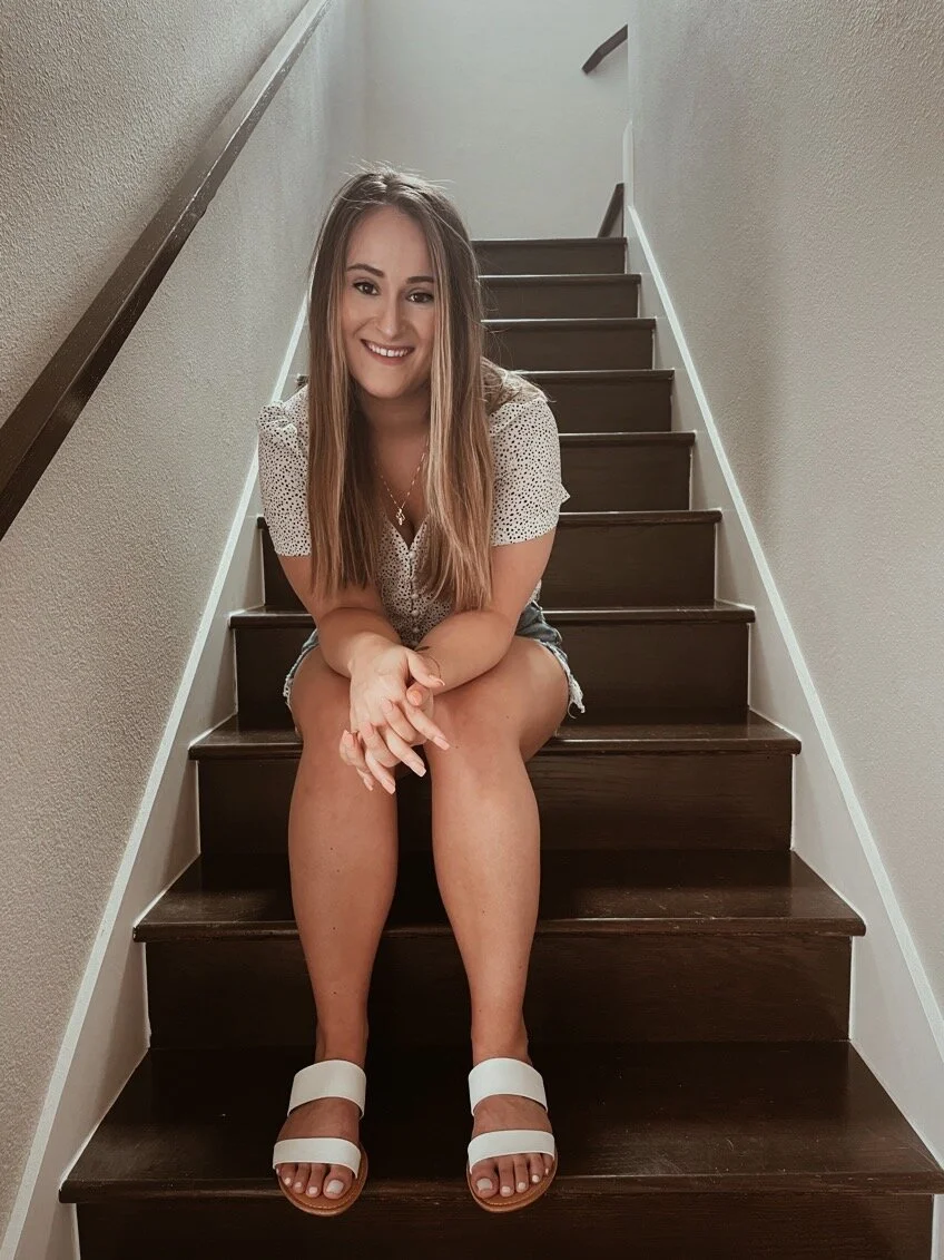 A young woman with long hair sitting on a staircase, smiling at the camera. She wears a white short-sleeved top, denim shorts, and white sandals.