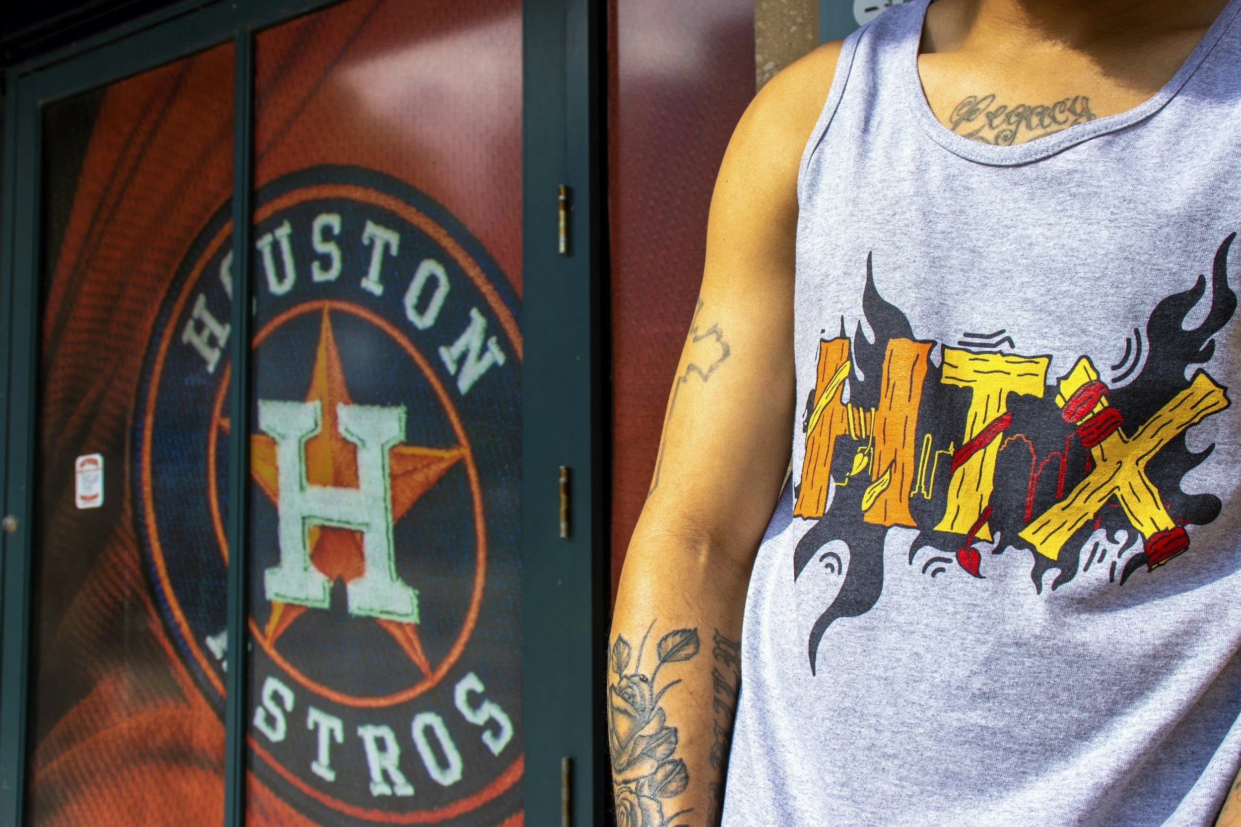 Close-up of a person's arm and shoulder with tattoos, wearing a sleeveless graphic T-shirt featuring a fiery skull, and standing in front of a U.S. Customs sign and door.