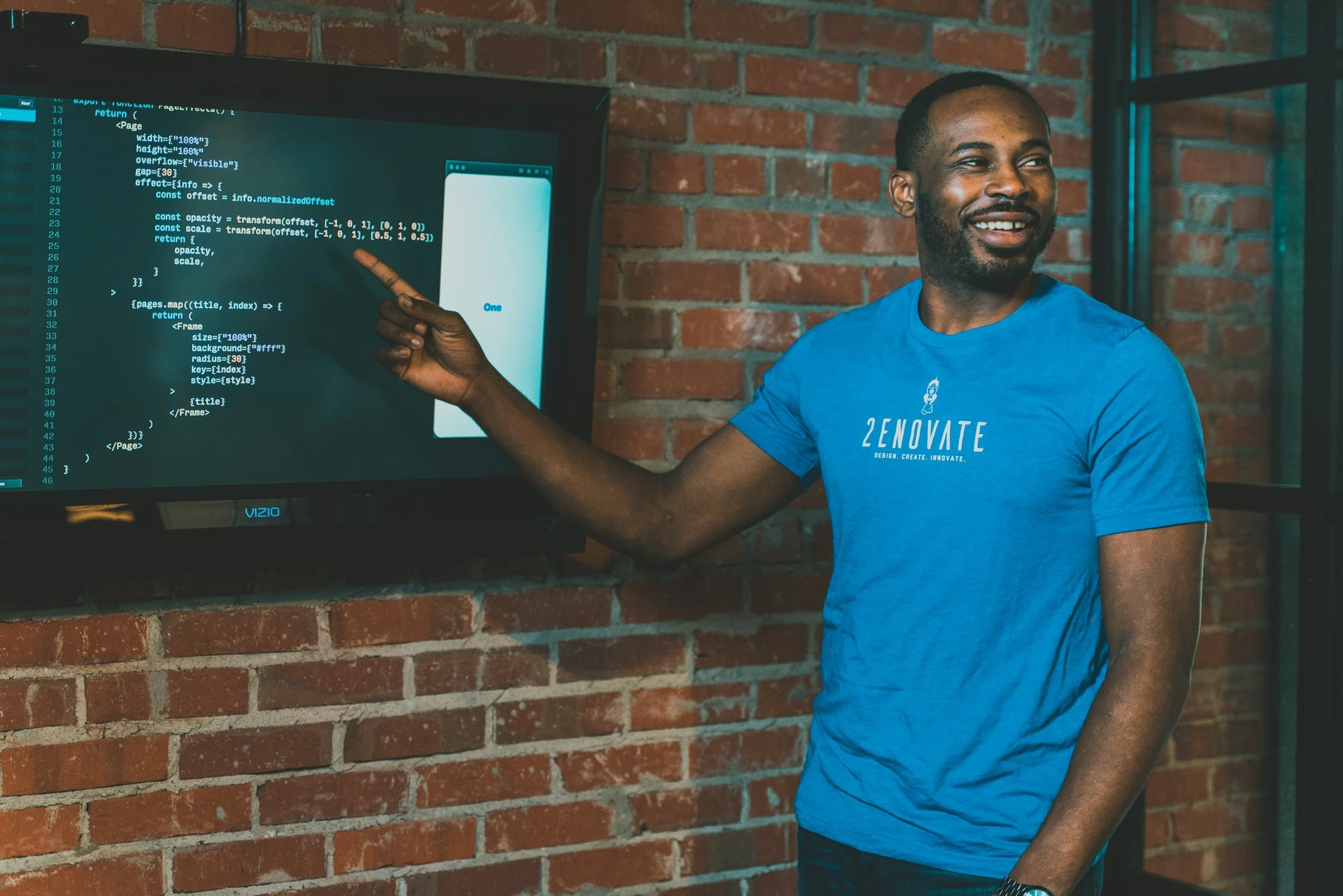 A man with a beard wearing a blue T-shirt pointing at a computer screen with coding on it, standing against a brick wall, smiling.