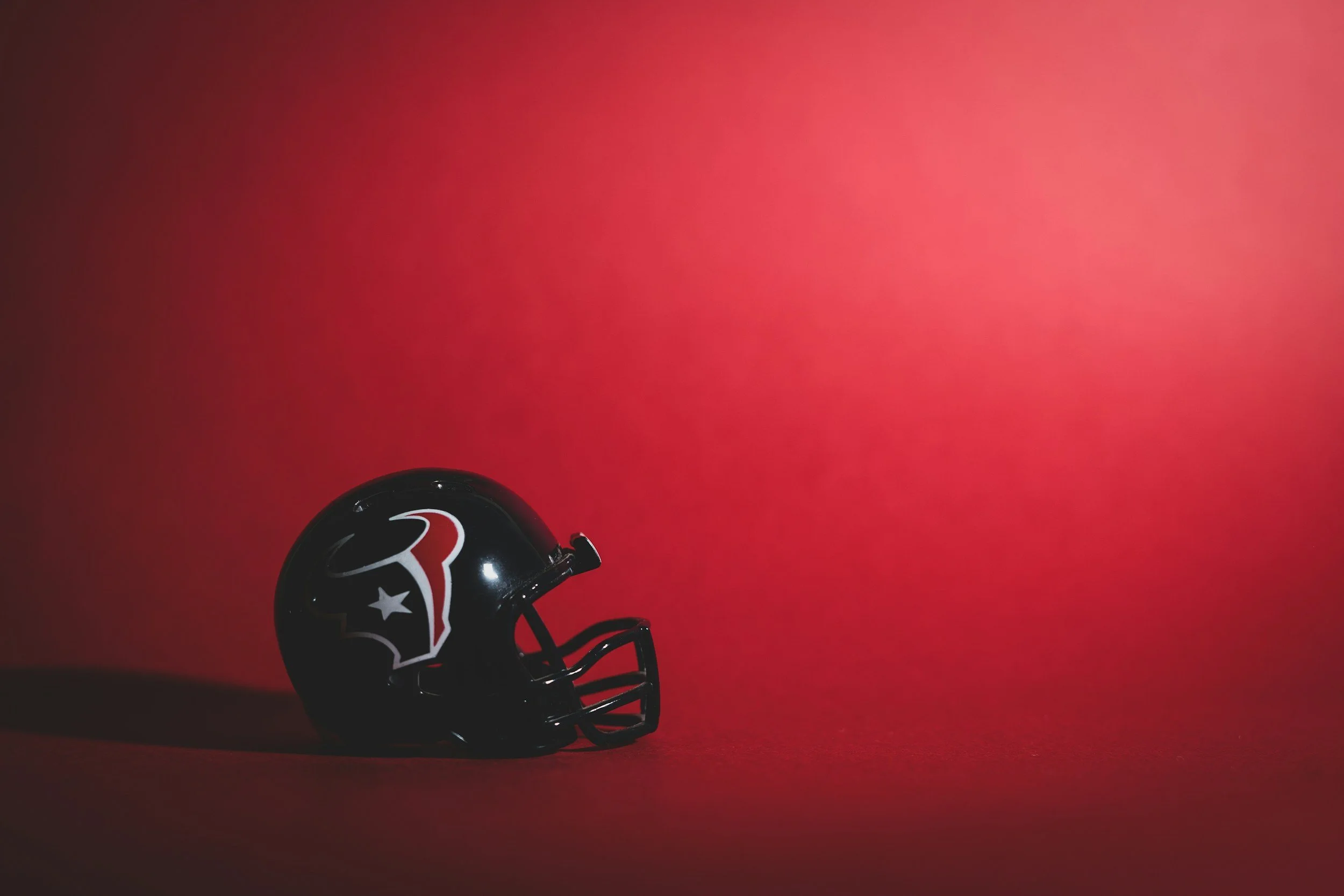 A small black NFL Houston Texans football helmet with a red, white, and silver logo, set against a solid red background.