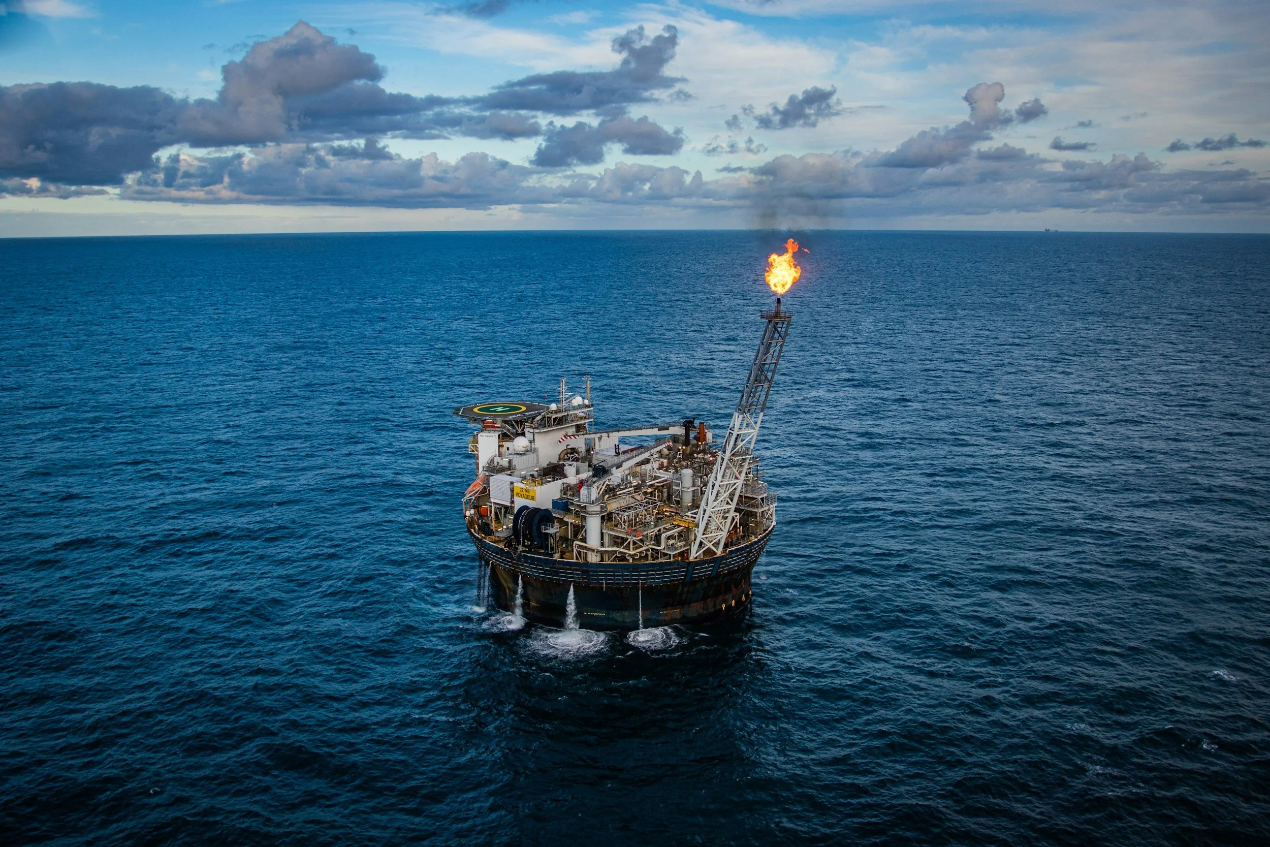 An offshore oil drilling platform in the ocean with a fire torch burning at the top, amid partly cloudy skies.