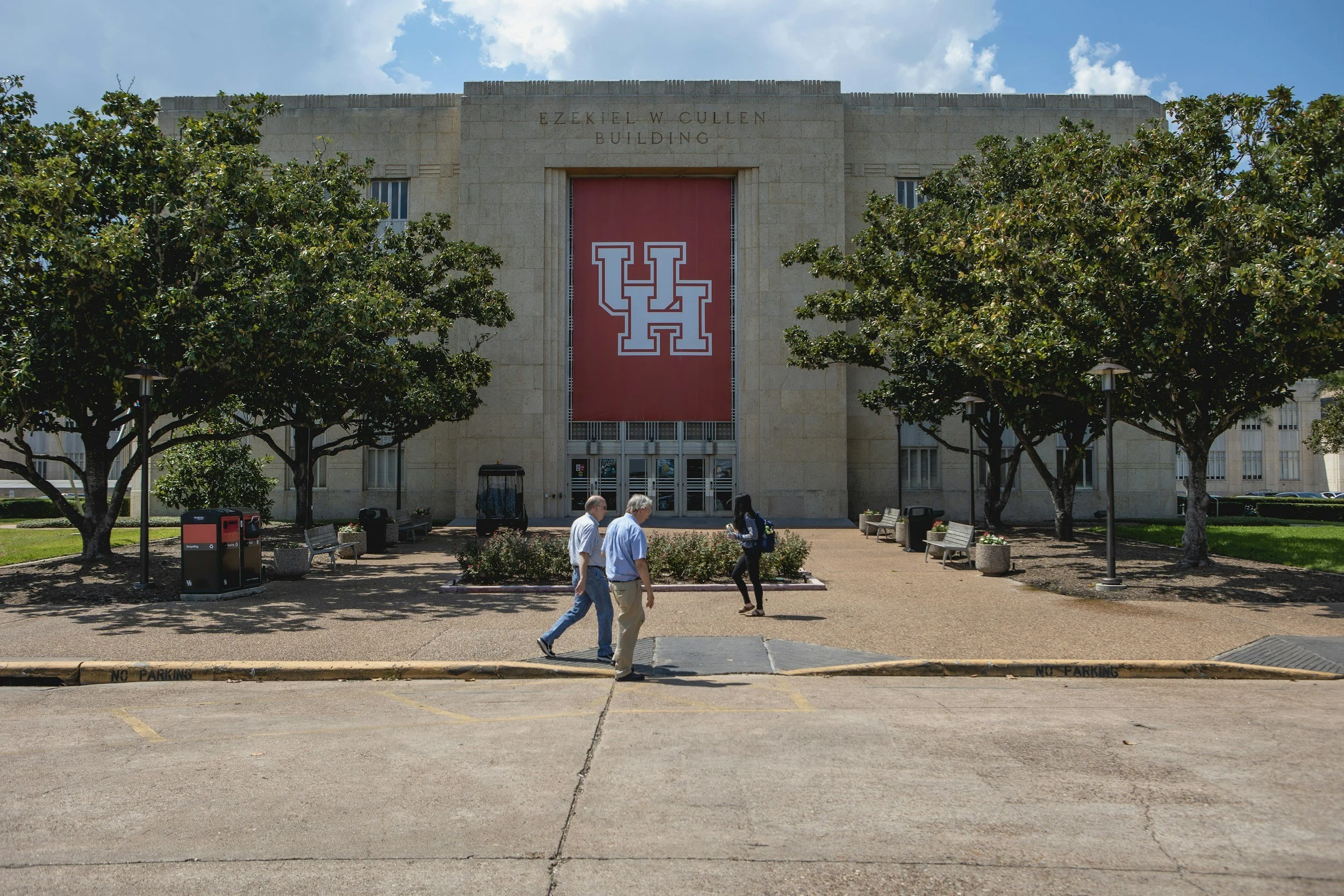 People walking on a sidewalk in front of a large building with trees lining the area. The building has a large red banner with the University of Houston logo and the name Ezekiel W. Cullen Building.