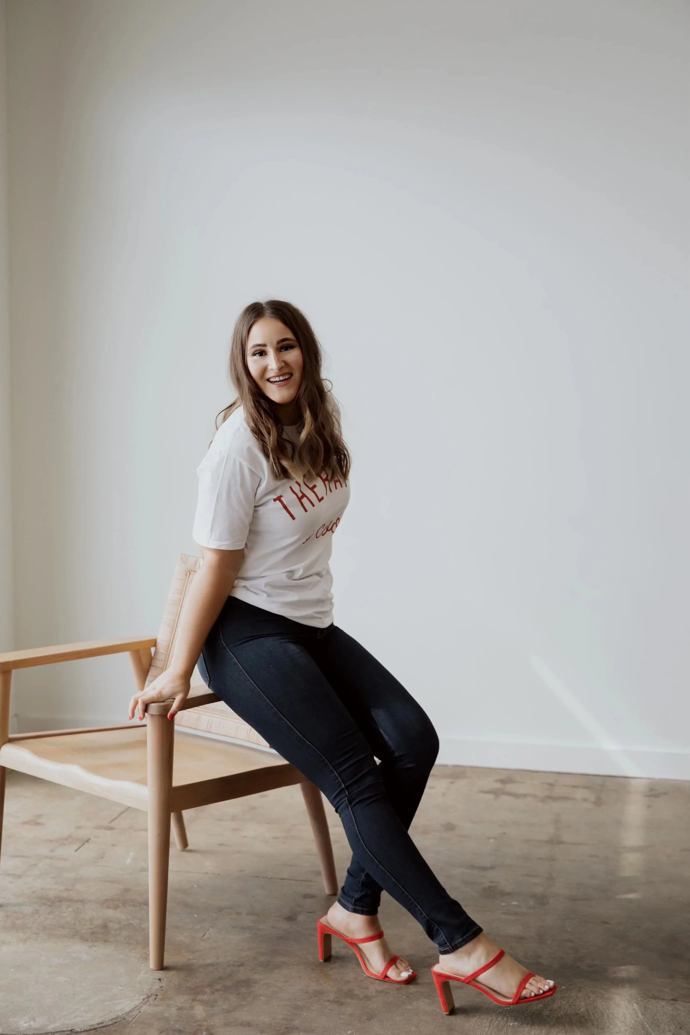 A woman sitting on a wooden chair in a room with a plain white wall, smiling at the camera, wearing a white T-shirt, dark jeans, and red high-heeled sandals.