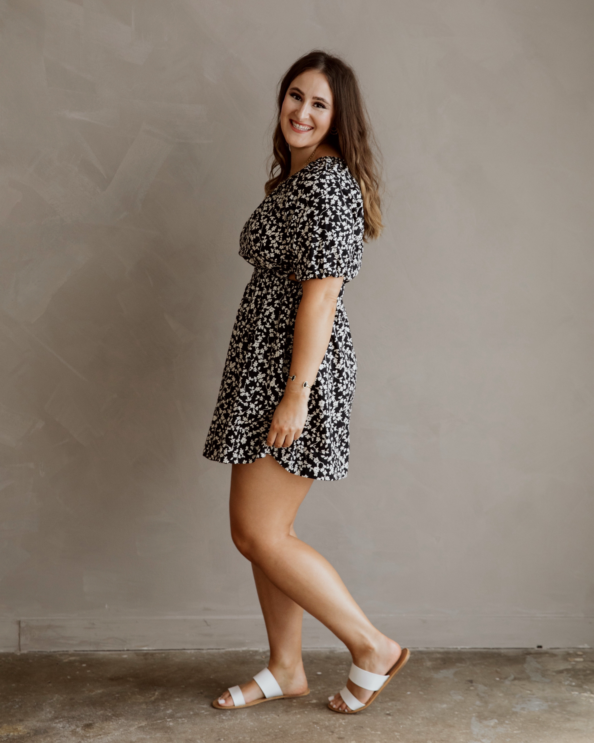 A woman with long wavy brown hair smiling and standing sideways against a neutral beige wall, wearing a black and white floral dress and white sandals.