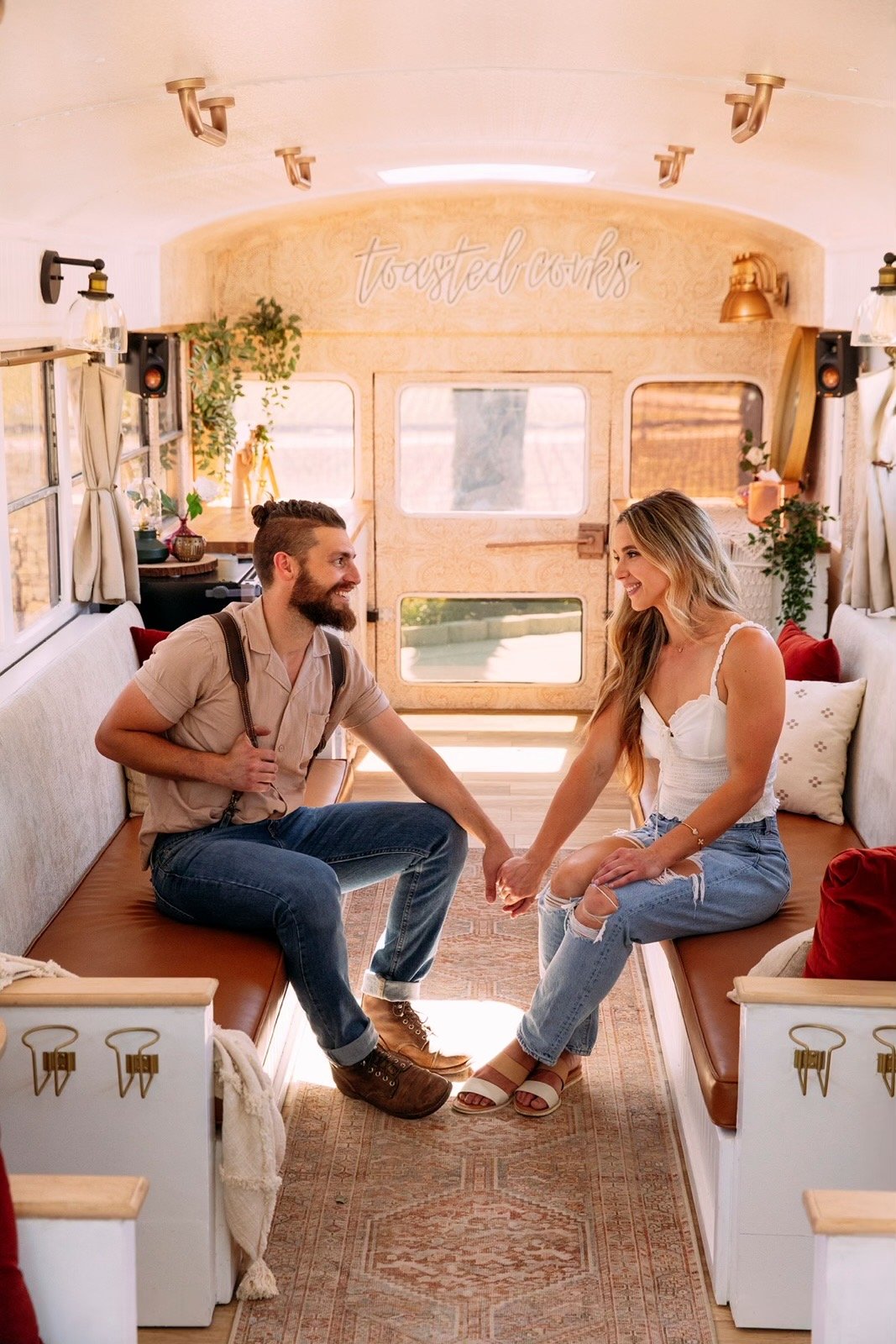 A couple sitting inside a converted bus with stylish decor, holding hands and smiling at each other. The interior features wooden accents, potted plants, cozy seating, and a sign that says 'toasted cooks.' The bus is bright and inviting.
