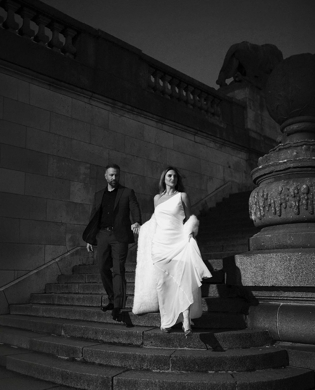 There is power in contrast. White against stone. Black against shadow. Movement against historic architecture. On the steps of Pont Alexandre III, Paris at night sharpens every detail. The gown flows effortlessly, the posture stays confident, the cit