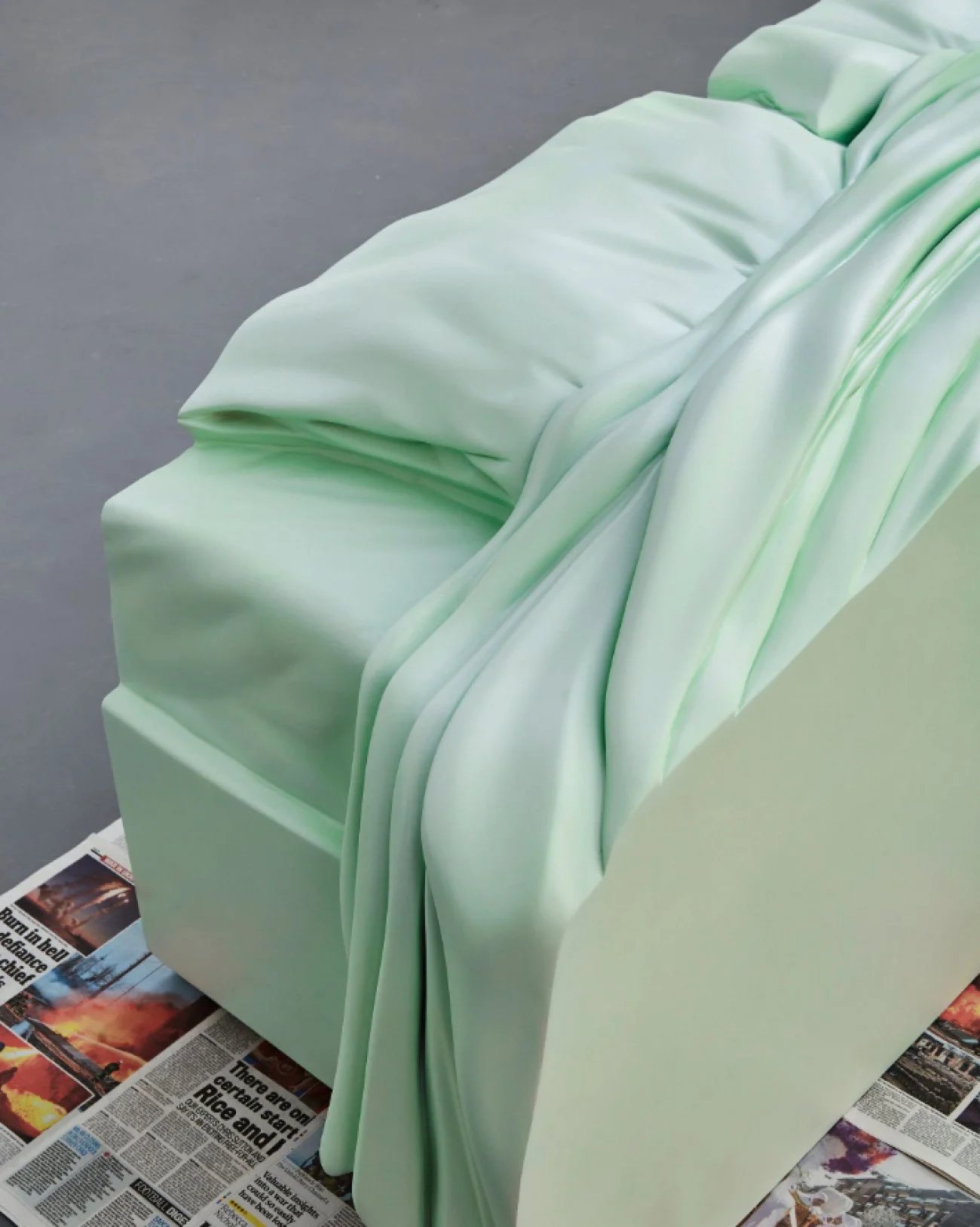 A large art installation resembles a sofa draped with flowing white fabric that has light green accents, positioned on a floor covered with newspapers.