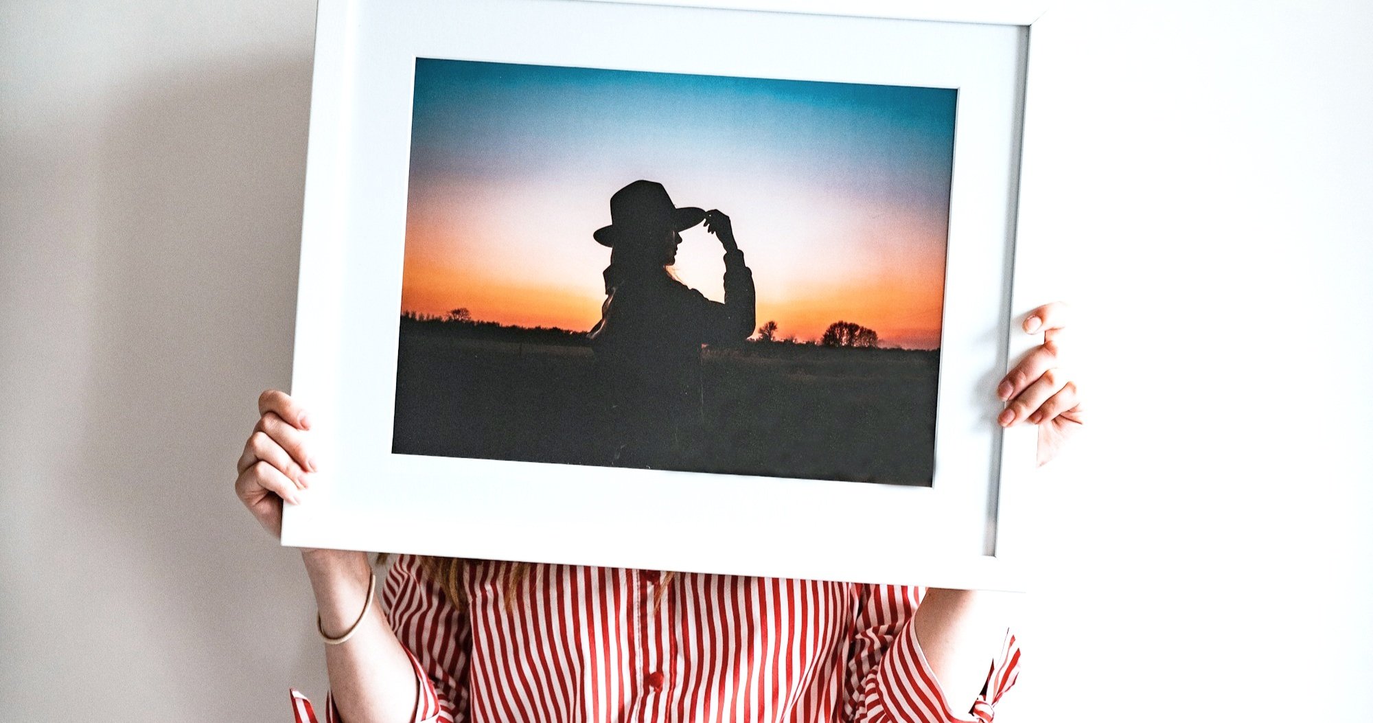 Person holding a framed photograph in front of their face. The photo shows a silhouette of a person wearing a hat at sunset, with warm orange and blue tones in the sky.