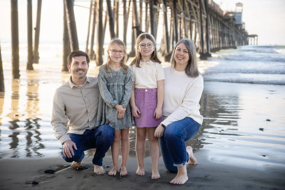 Family of four photographed barefoot on the beach near a pier
