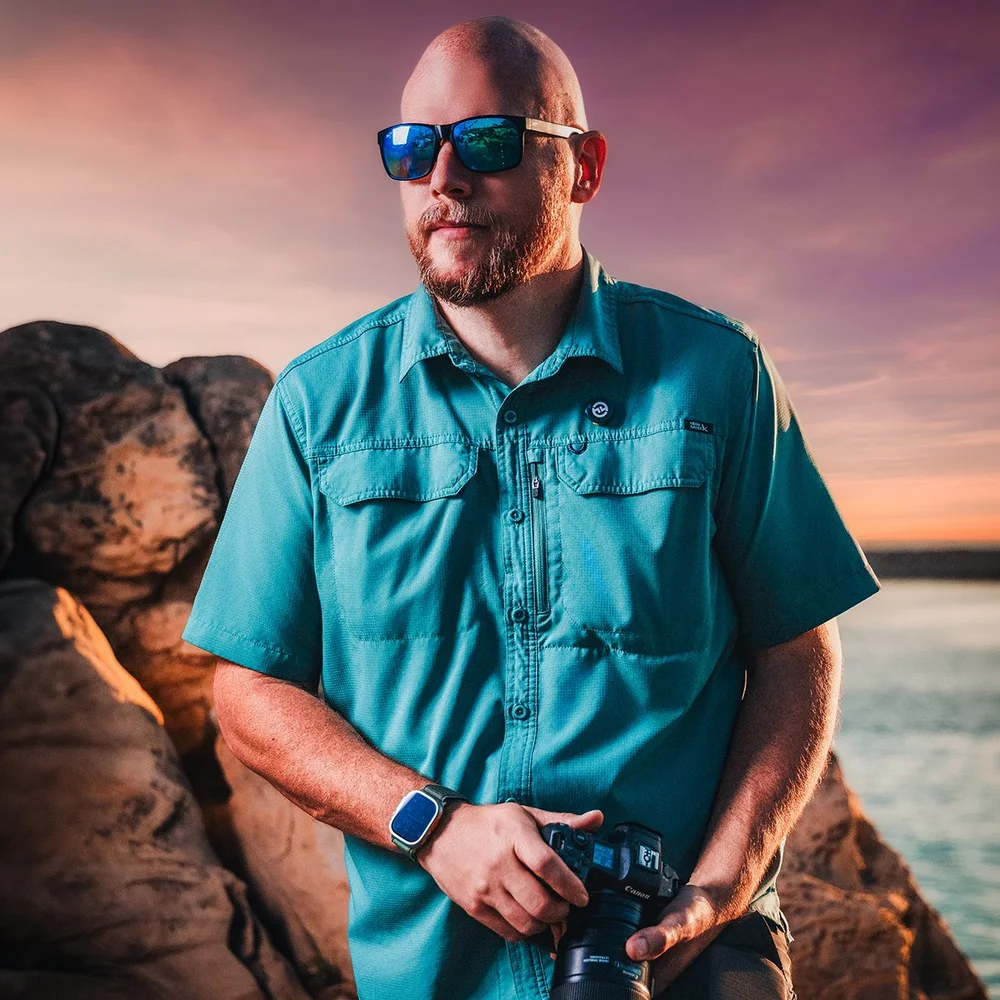 Joseph holding a camera on the rocks at sunset by the water