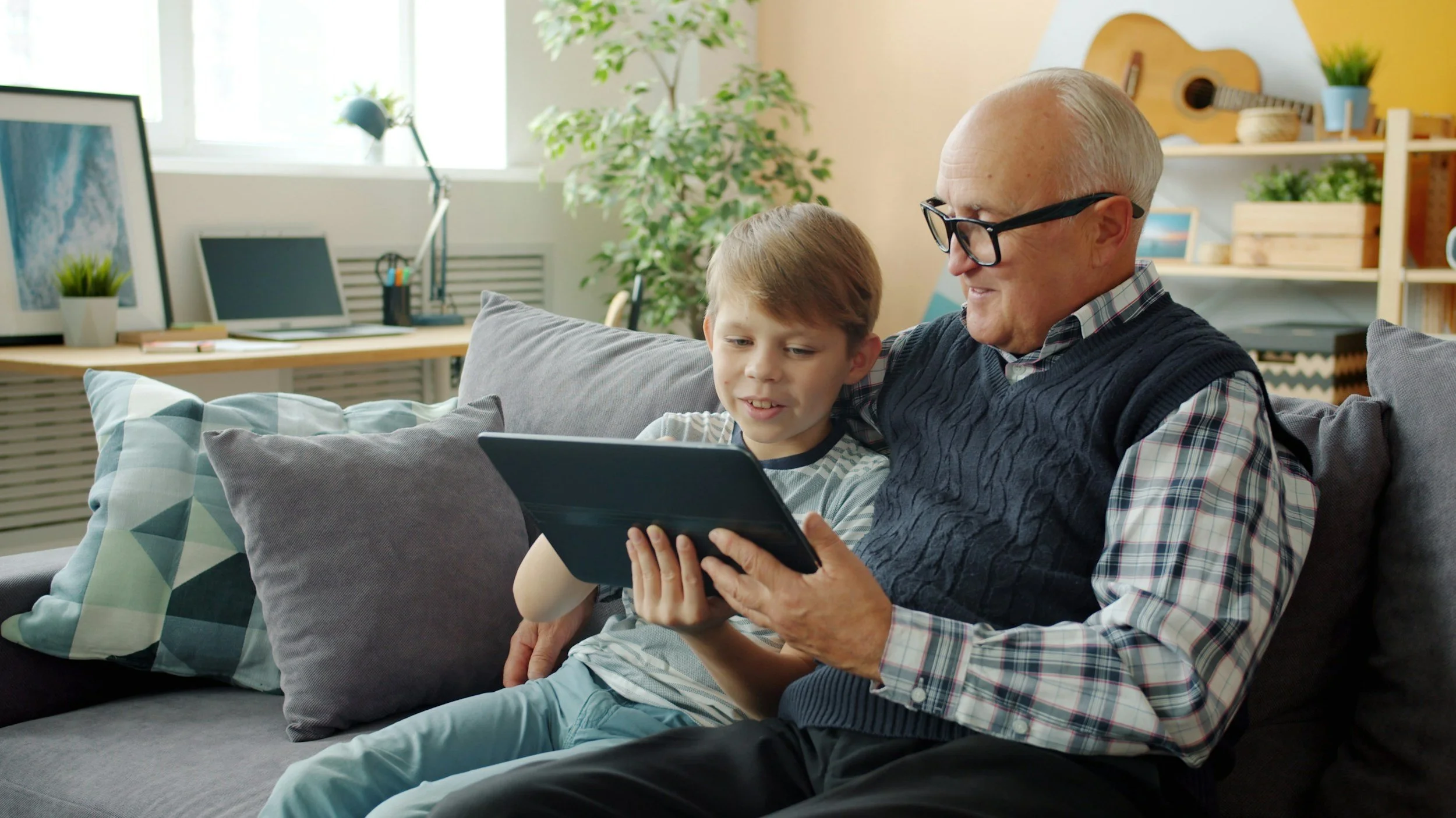 An elderly man and a young boy sitting on a couch, looking at a tablet together in a cozy living room with plants and musical instruments in the background.
