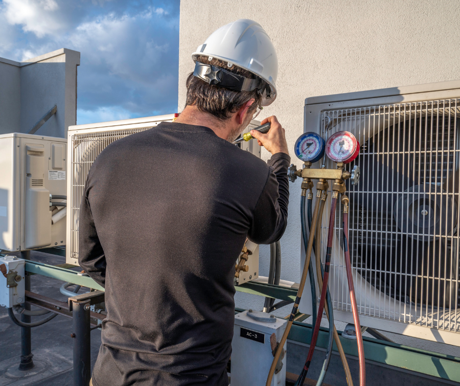 A technician wearing a white safety helmet and black shirt is working on an air conditioning unit outside a building, using tools to check the system.