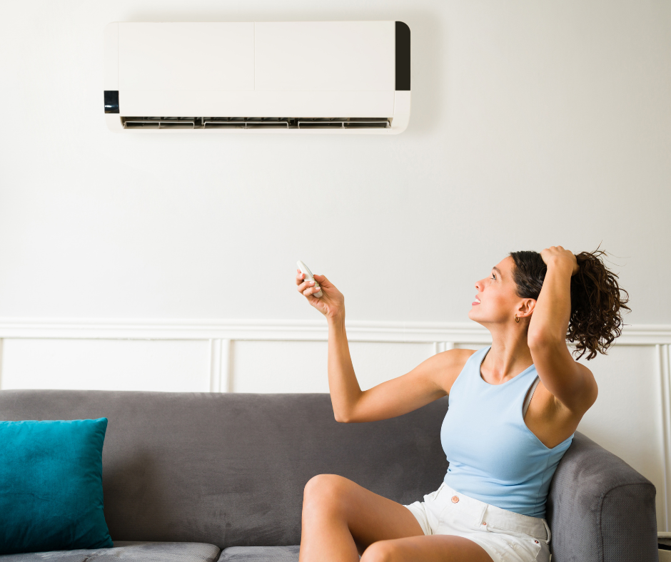 A woman sitting on a gray sofa, holding a remote control, smiling and adjusting her hair while looking at a wall-mounted air conditioner.