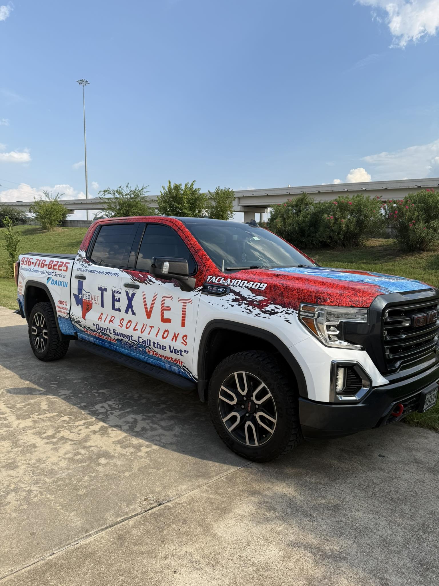 A white pickup truck with Texas Vet Air Solutions branding on the side, parked on a concrete surface under a clear blue sky with some clouds. There are green bushes and a highway overpass in the background.