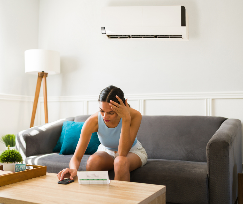 Woman sitting on gray couch in living room, looking stressed, with a calculator and papers on coffee table, air conditioner on wall, and lamp in background.