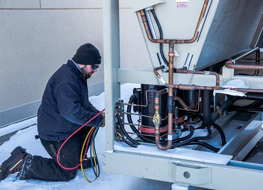 Technician working on HVAC unit outside in snowy weather, inspecting copper piping and electrical components.