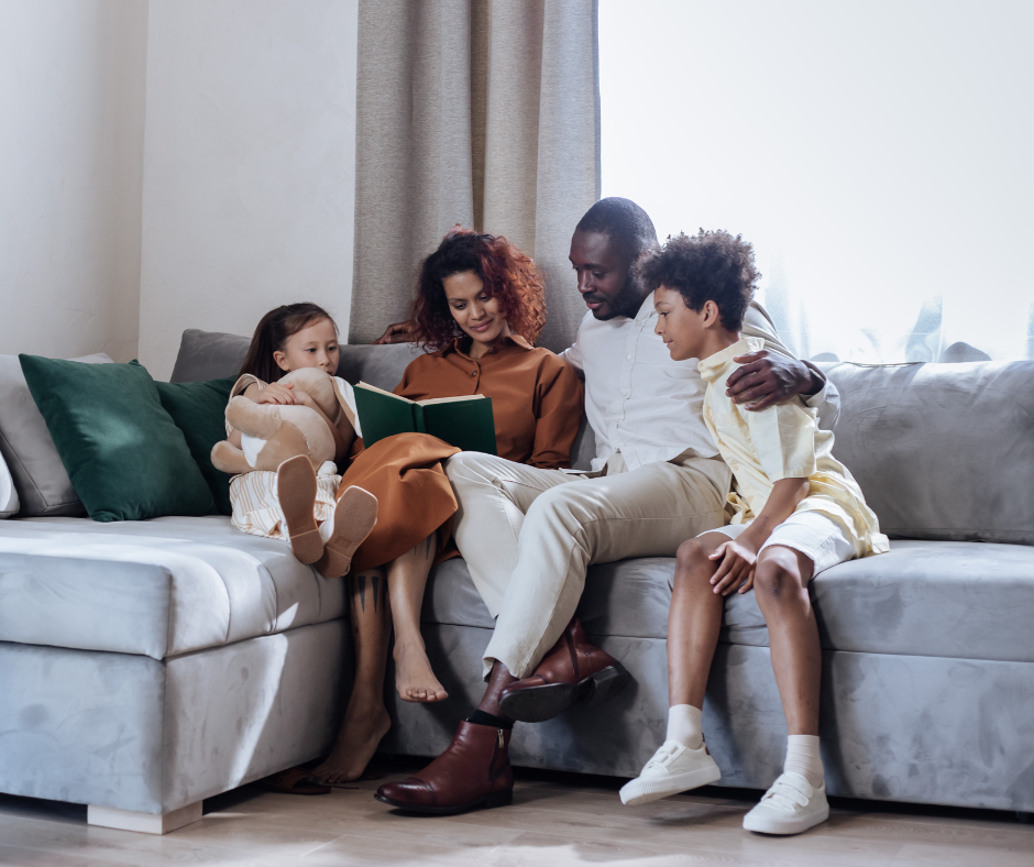 Family of four sitting on a gray sofa, reading a book together in a living room with beige walls and curtains.