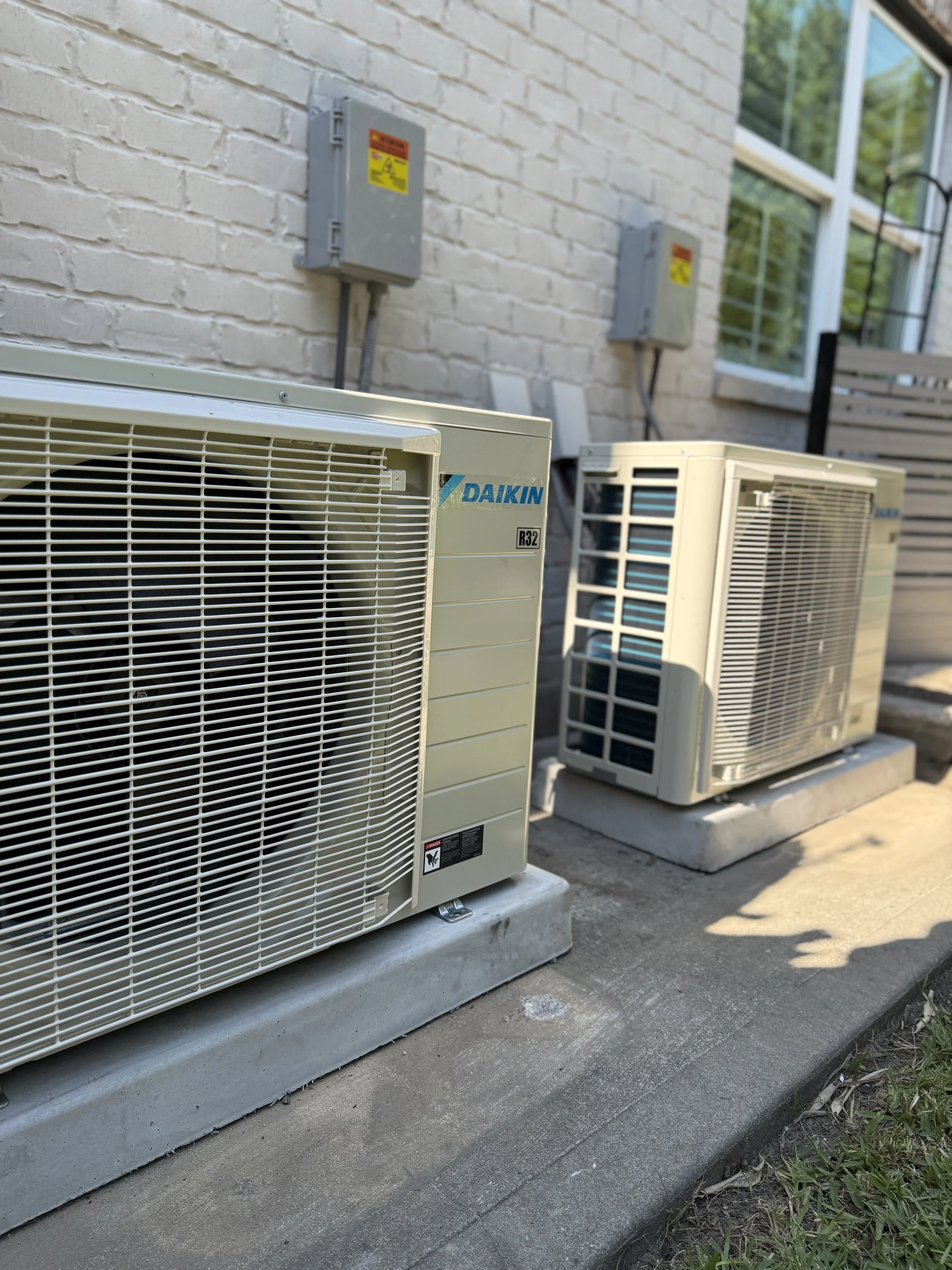 Three outdoor air conditioning units installed outside a building with a brick wall. The units are on concrete pads and are connected to electrical panels mounted on the wall. The scene is sunny, and there is some greenery at the edge of the image.