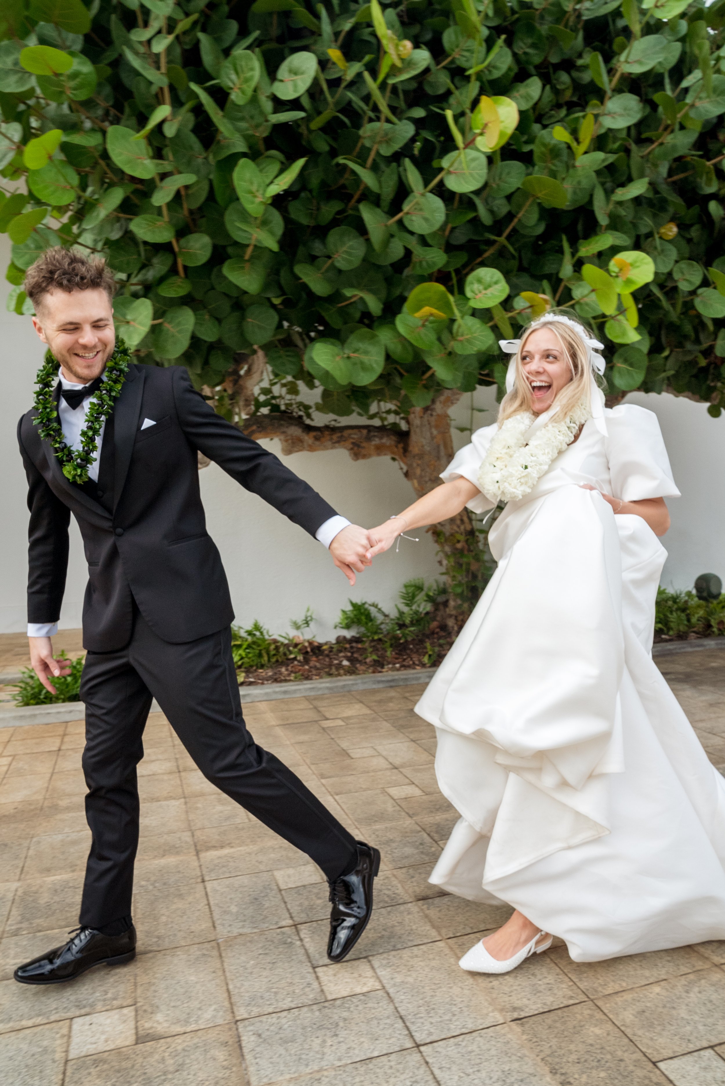 A bride and groom dancing happily outdoors on a checkered tile patio with a leafy tree in the background. The groom is wearing a black tuxedo with a lei necklace, and the bride is in a white wedding dress with a floral lei and a big smile.Laie Temple