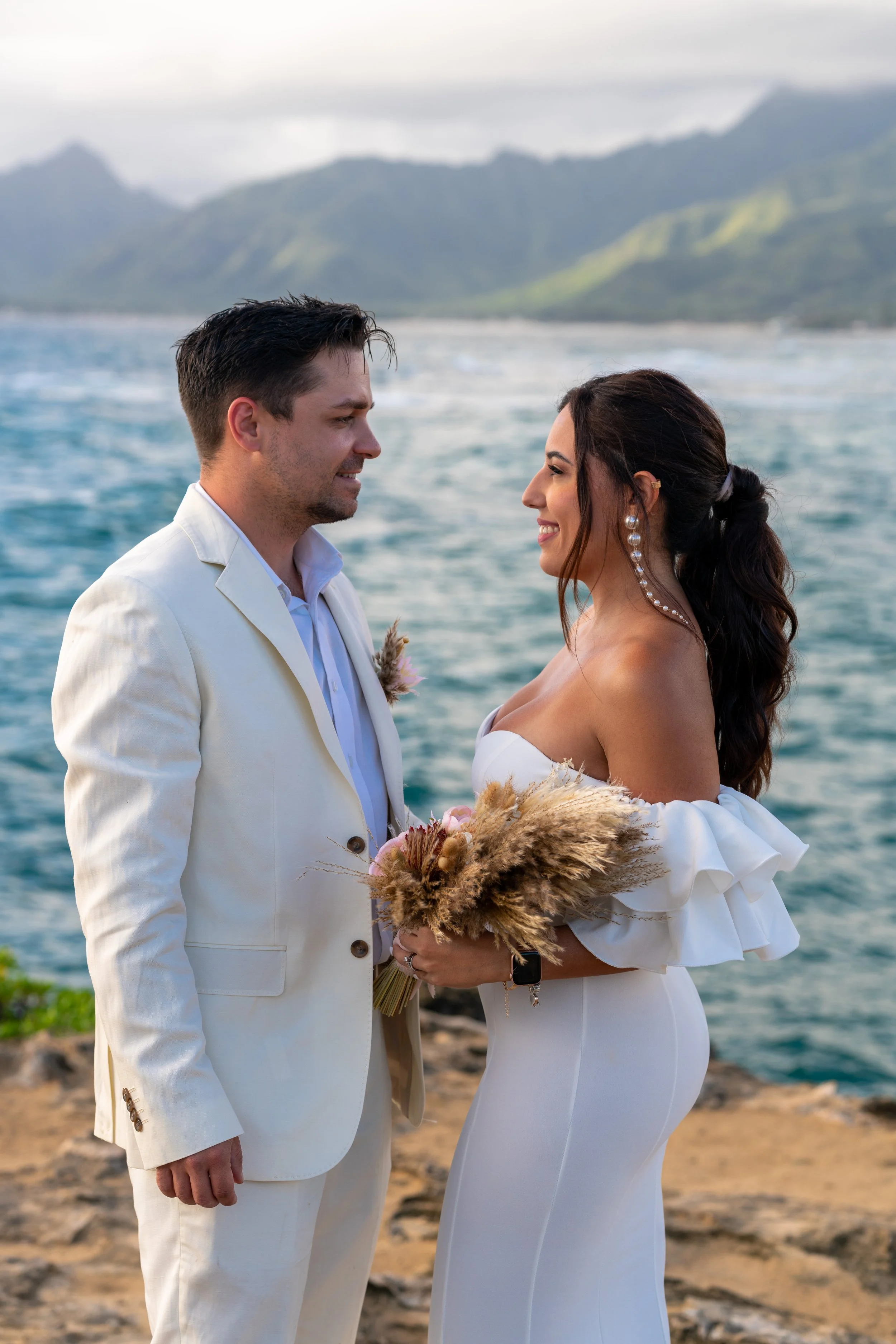 A bride and groom stand facing each other on a rocky shoreline by the ocean, with mountains in the background during their wedding ceremony.