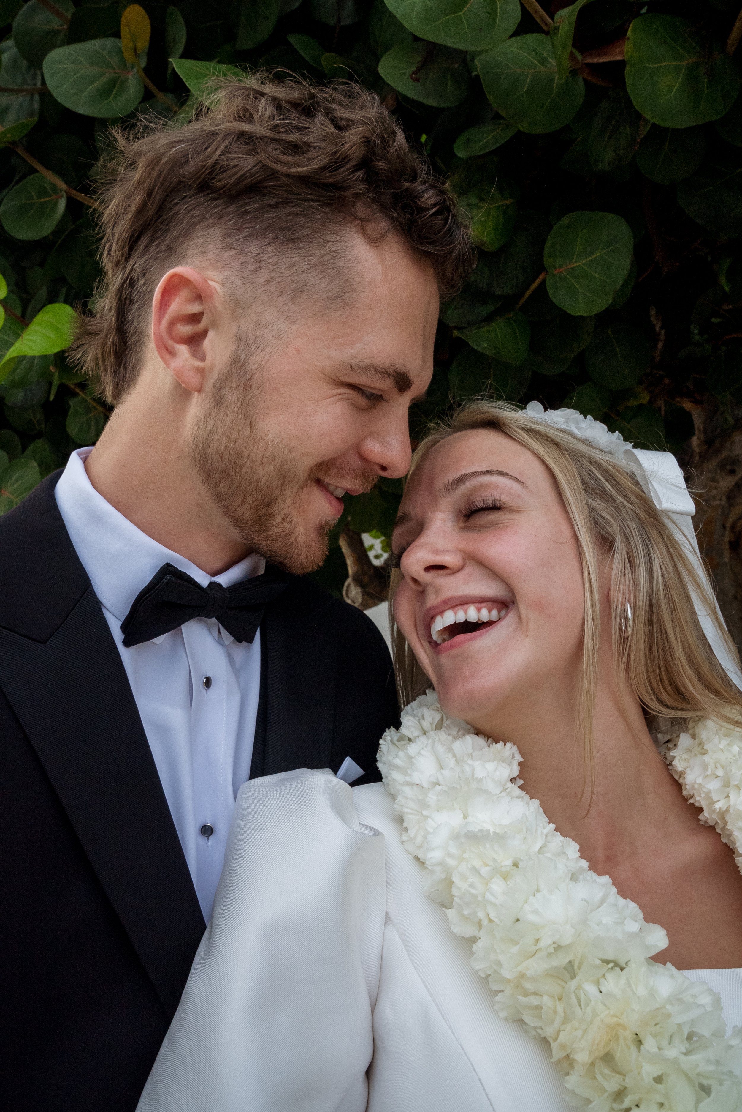 A smiling bride and groom on their wedding day, standing close together outdoors with green leaves in the background in Laie Hawaii.