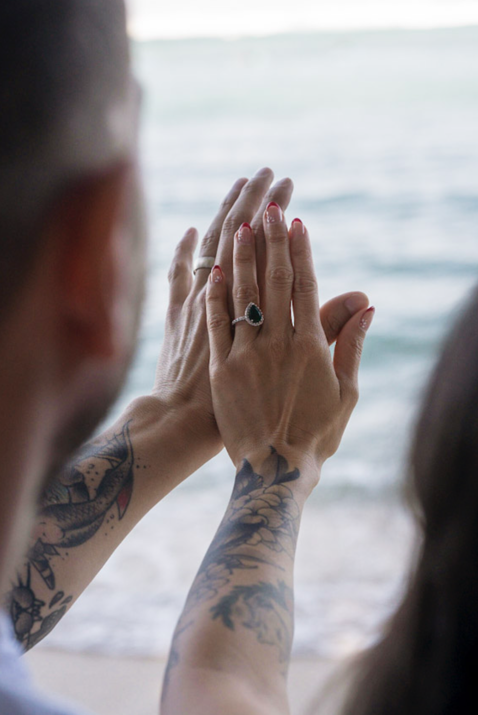 Hands touching with a ring on finger, tattoos visible on arms, near a body of water, possibly at a beach.