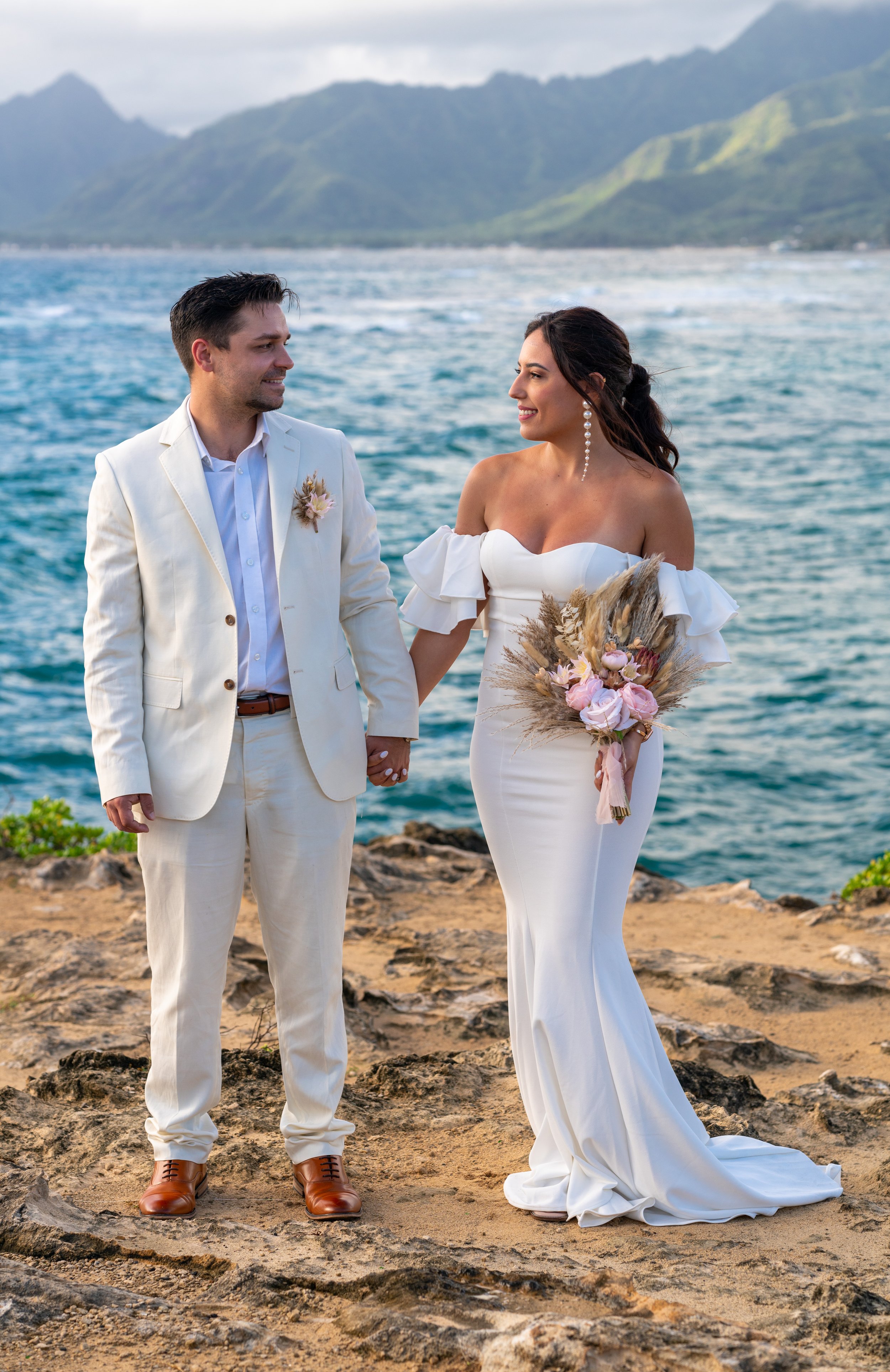 A bride and groom holding hands in front of the ocean and mountains, dressed in wedding attire, wedding ceremony on a Hawaii beach.